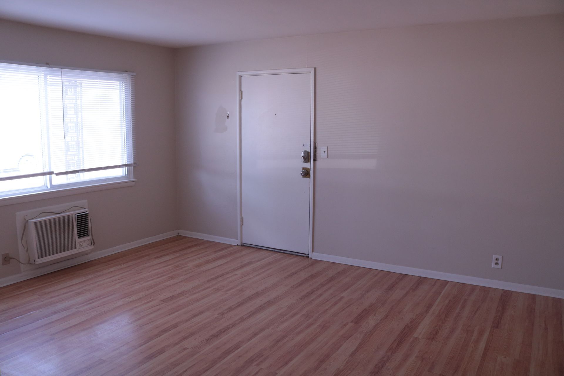 Empty room with wood floors, a window with blinds, and a closed door. Wall-mounted air conditioner.