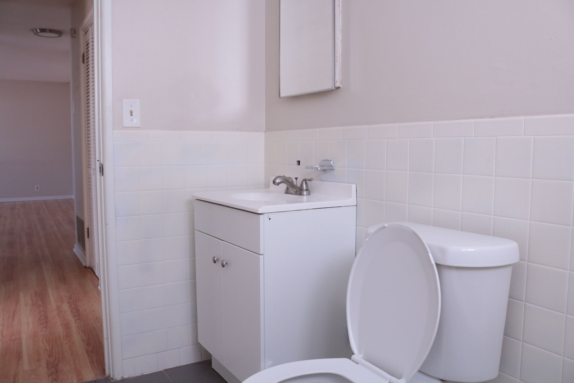 White bathroom with toilet, sink, and cabinet. Tile wainscoting and neutral wall paint.
