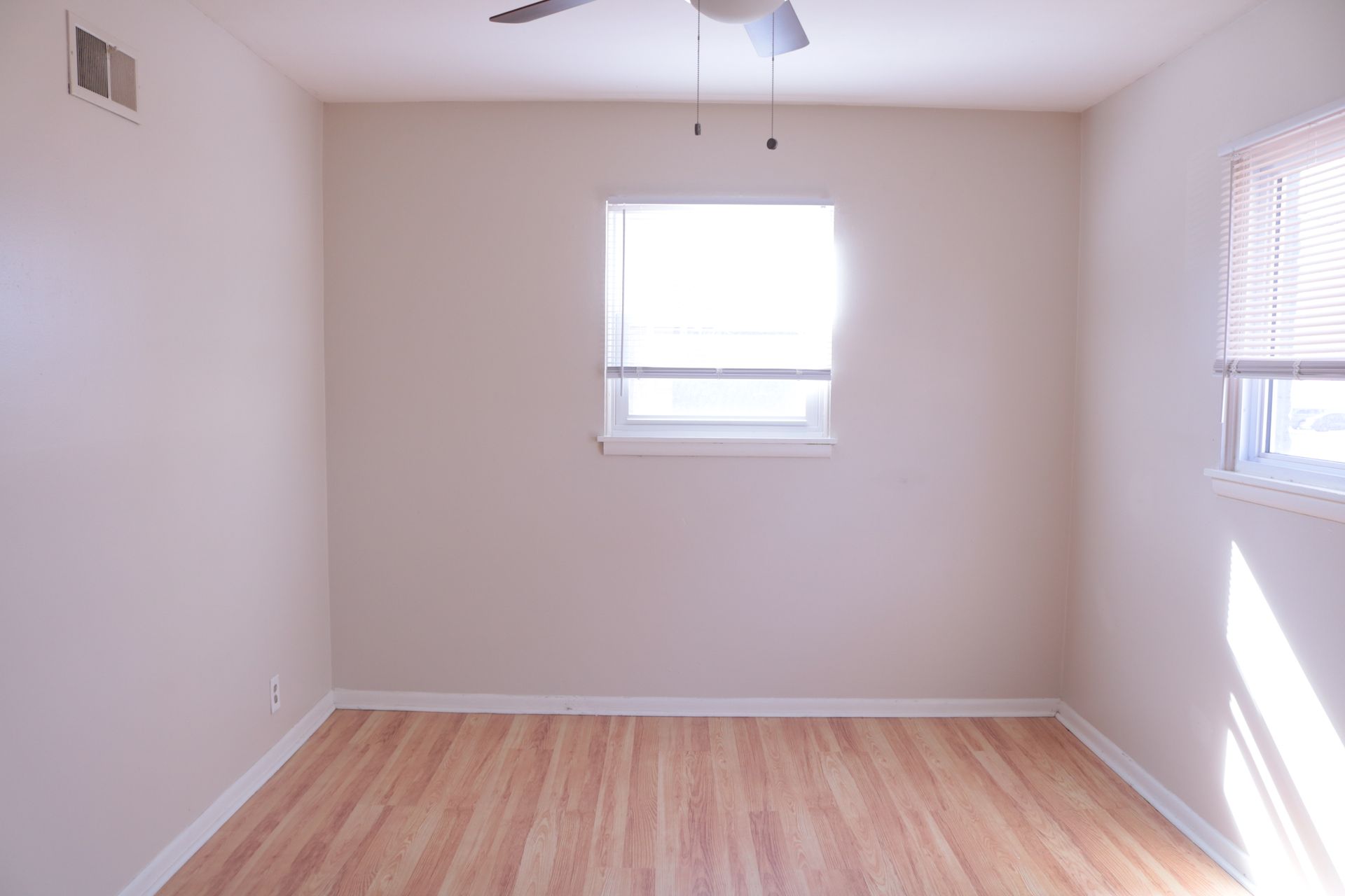 Empty room with hardwood floors, two windows with blinds, and a ceiling fan.