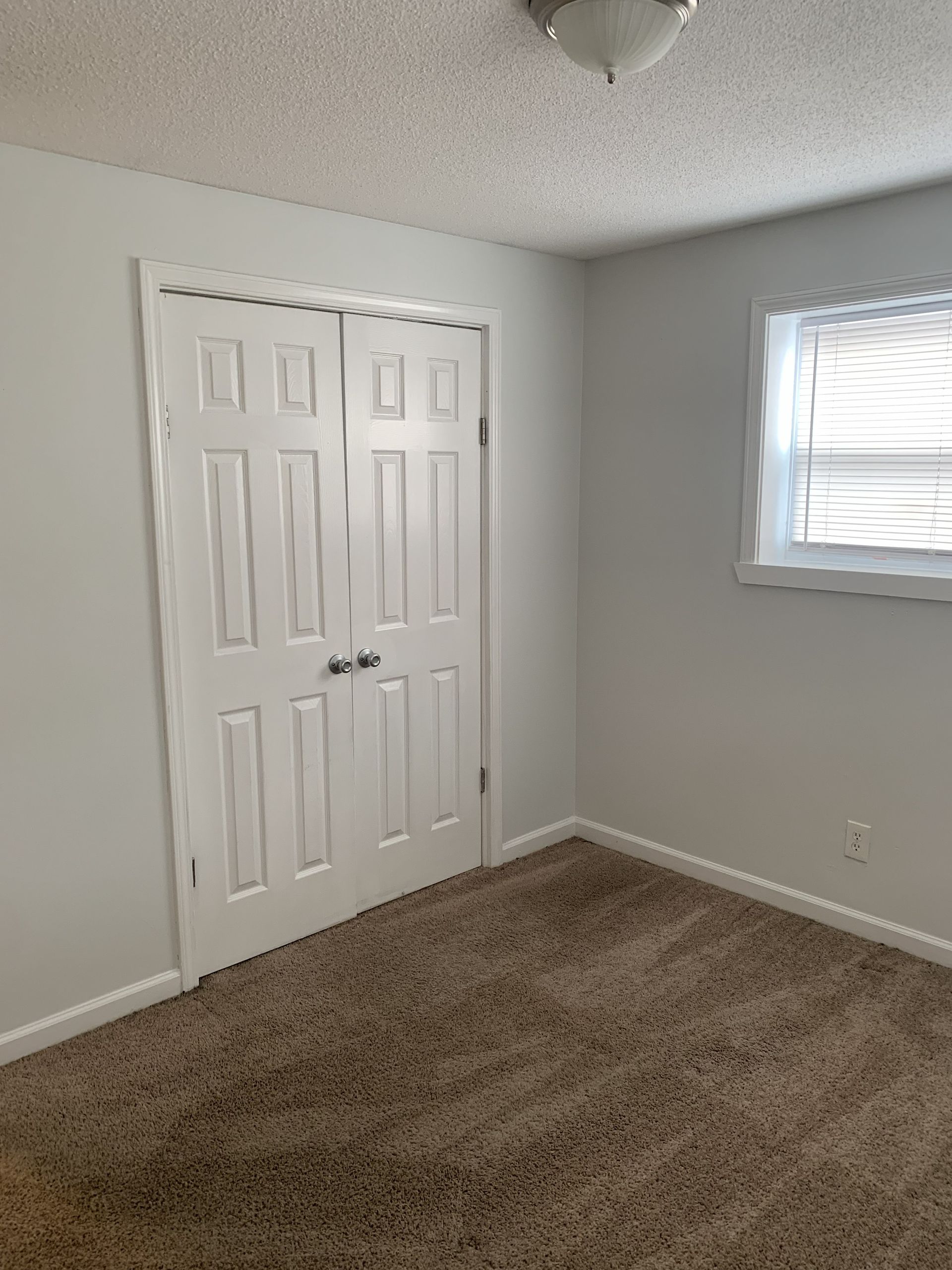 Interior view of a small room with a closet, window, light fixture, and brown carpet. The walls are white.