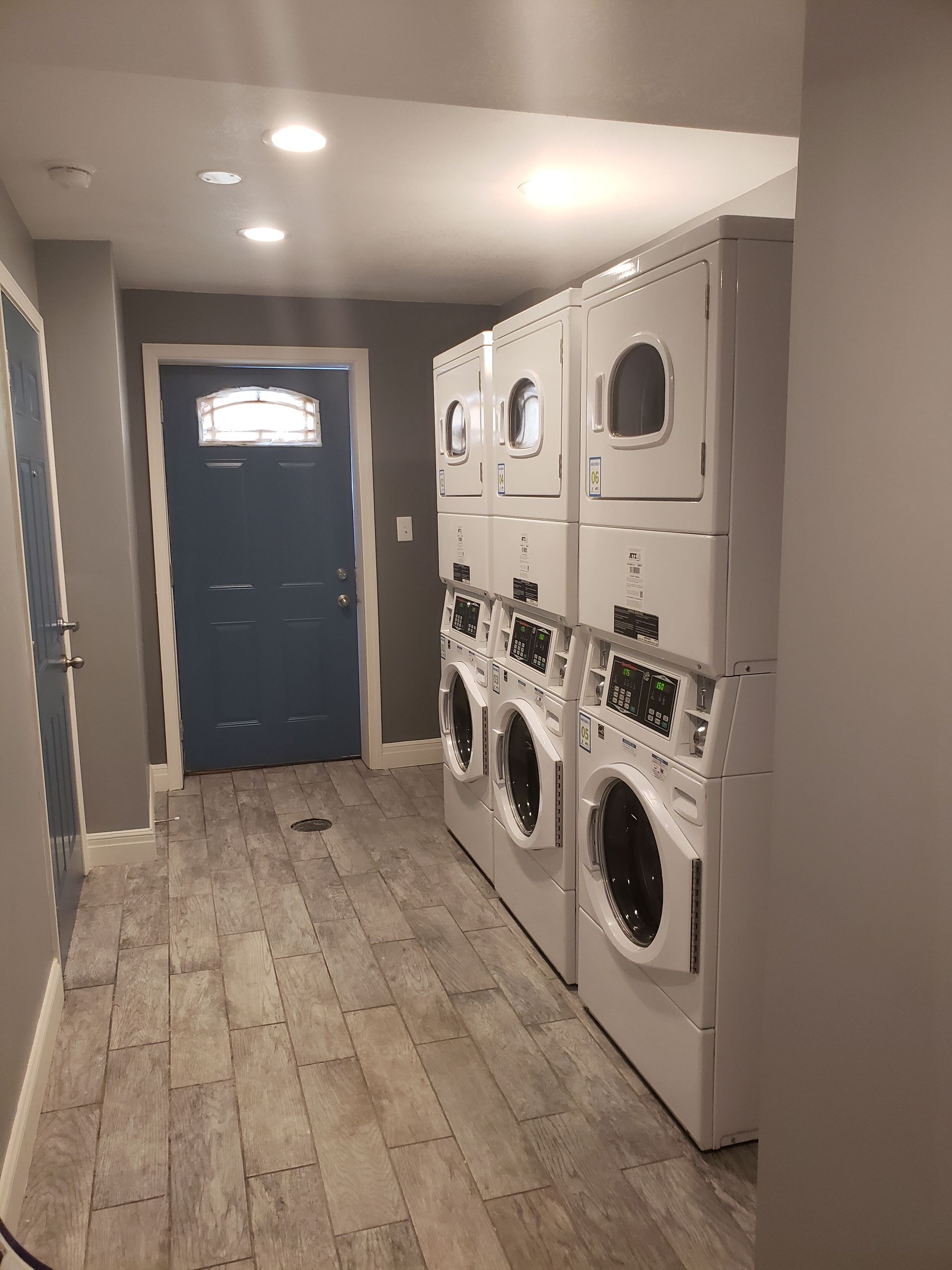 Laundry room with several stacked washers and dryers. Blue door at the end of the hallway. Light wood flooring.