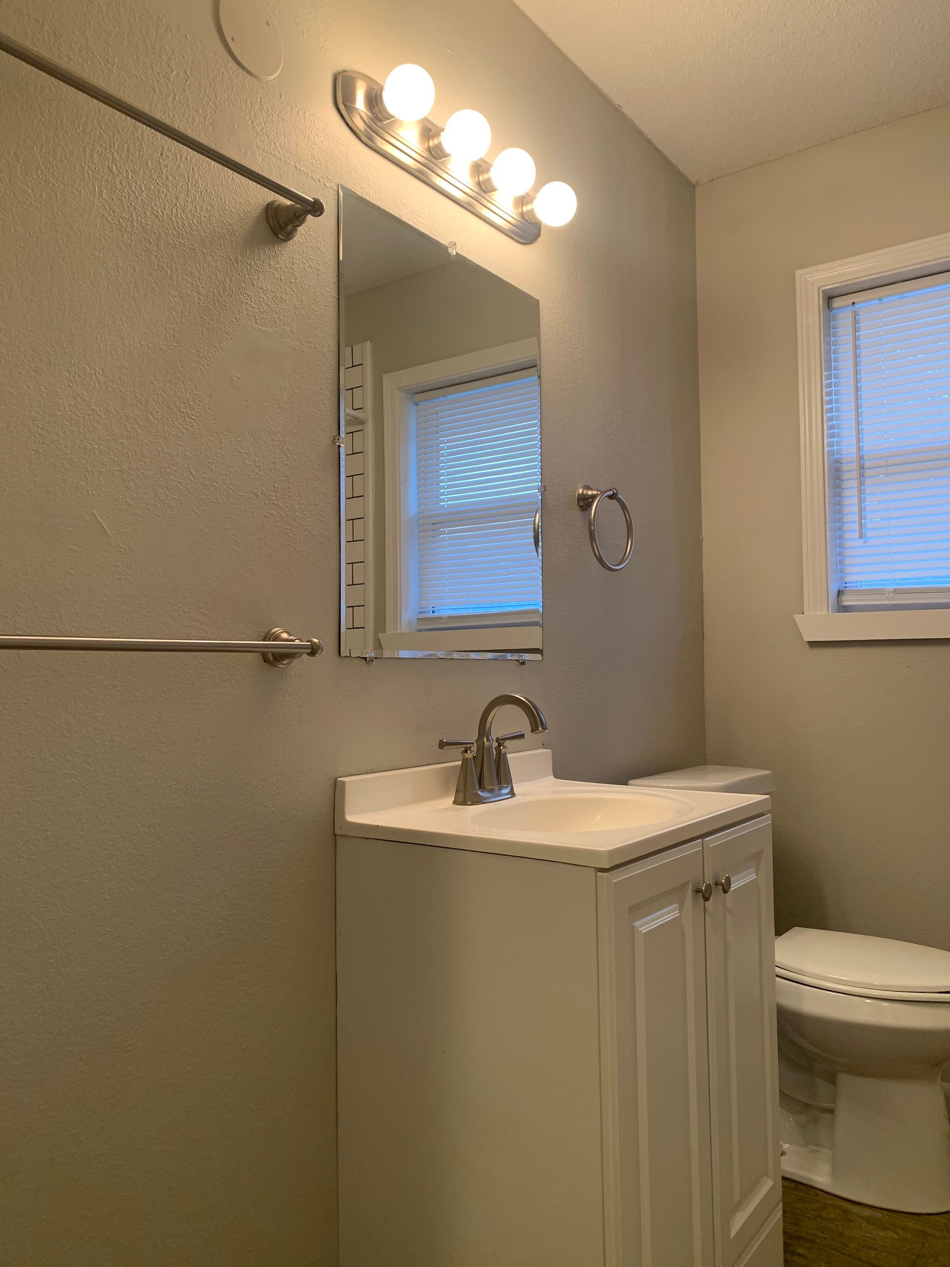 Bathroom with white vanity, mirror, lights, and toilet, gray walls, and blinds.