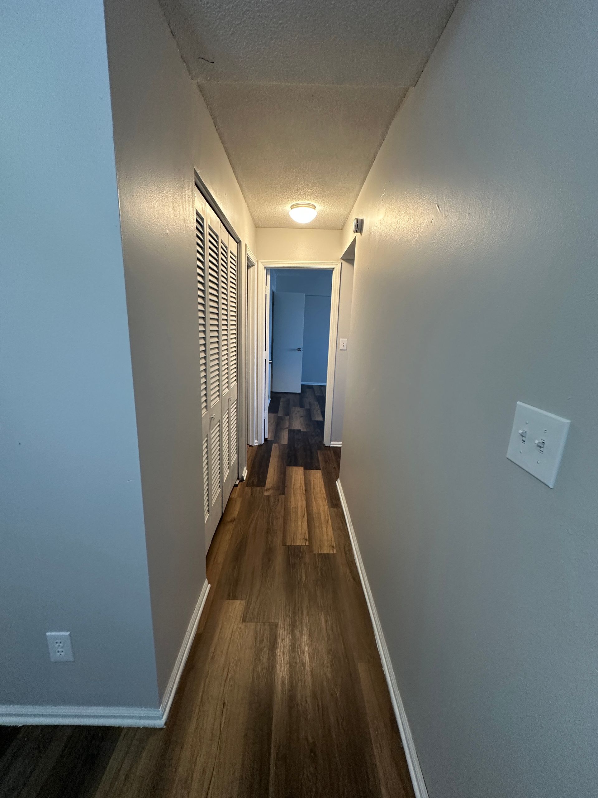 Narrow hallway with wooden floor, white closet doors, and light gray walls, leading to a doorway.