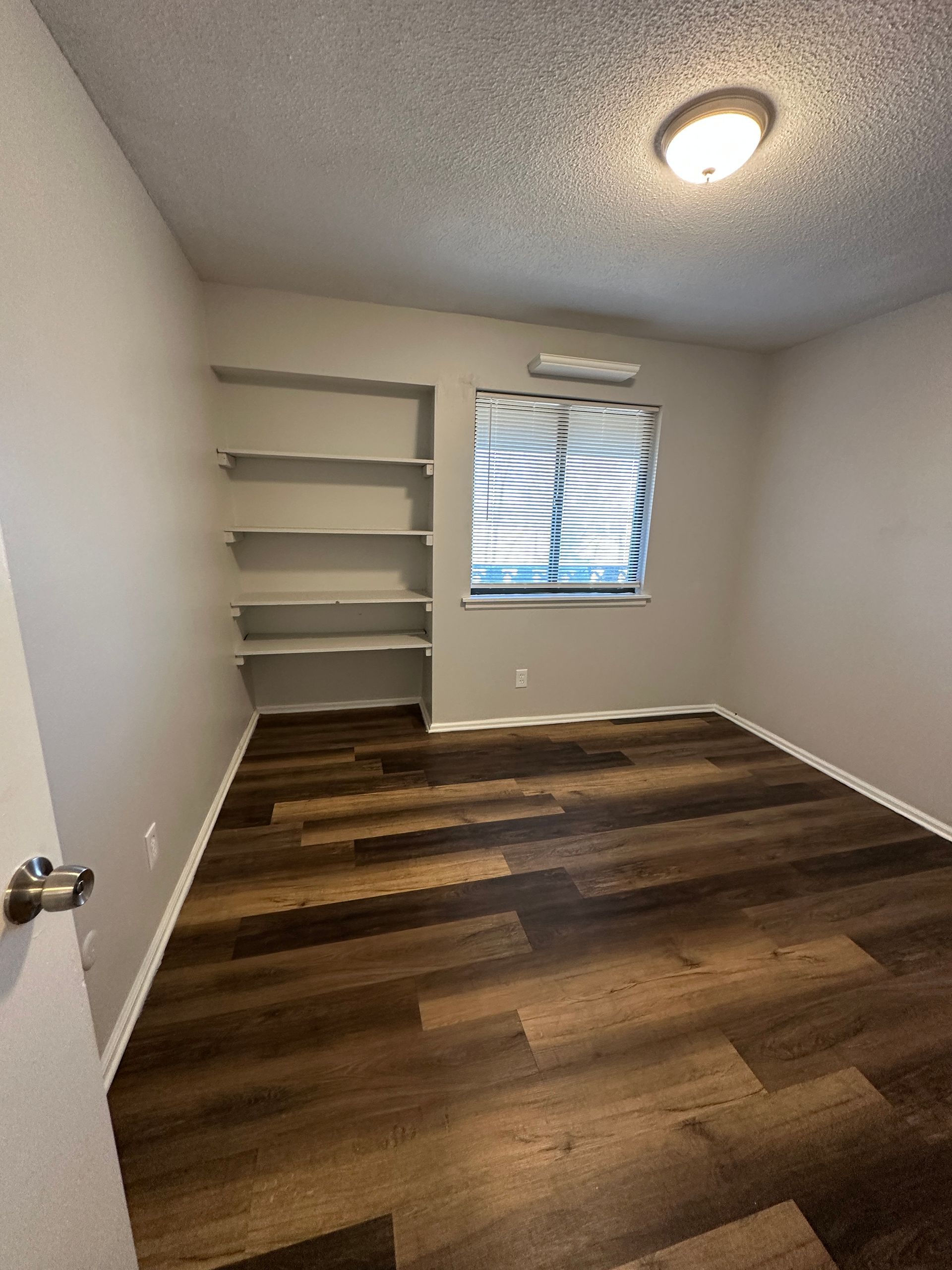 Empty room with built-in shelves, a window, a dark wood-look floor, light-colored walls, and ceiling light.