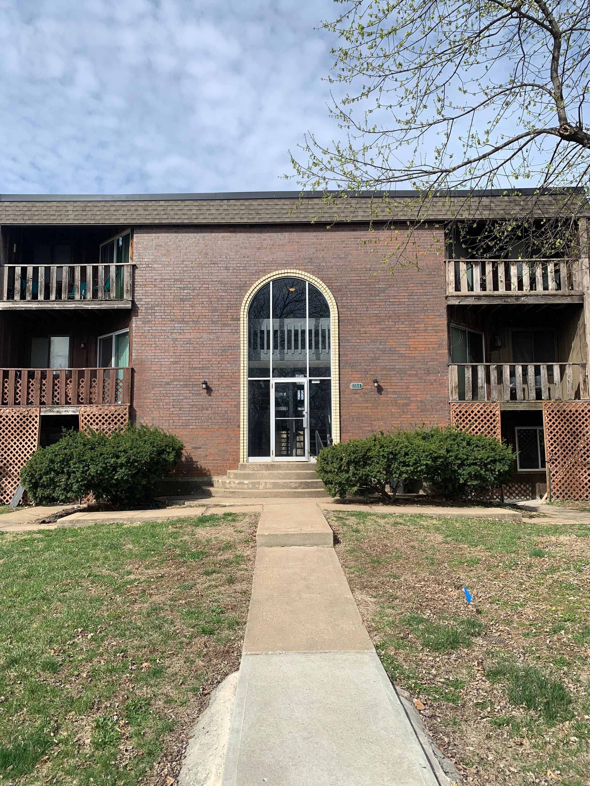 Apartment building with brick facade and arched entrance. A concrete path leads to the front door, with shrubbery and trees.