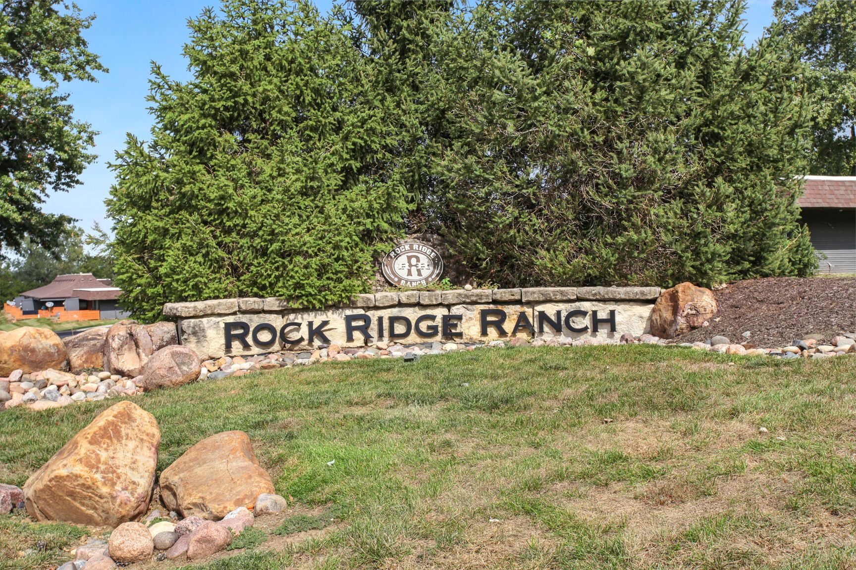 Rock Ridge Ranch sign in a grassy area with large rocks and green bushes against a blue sky.