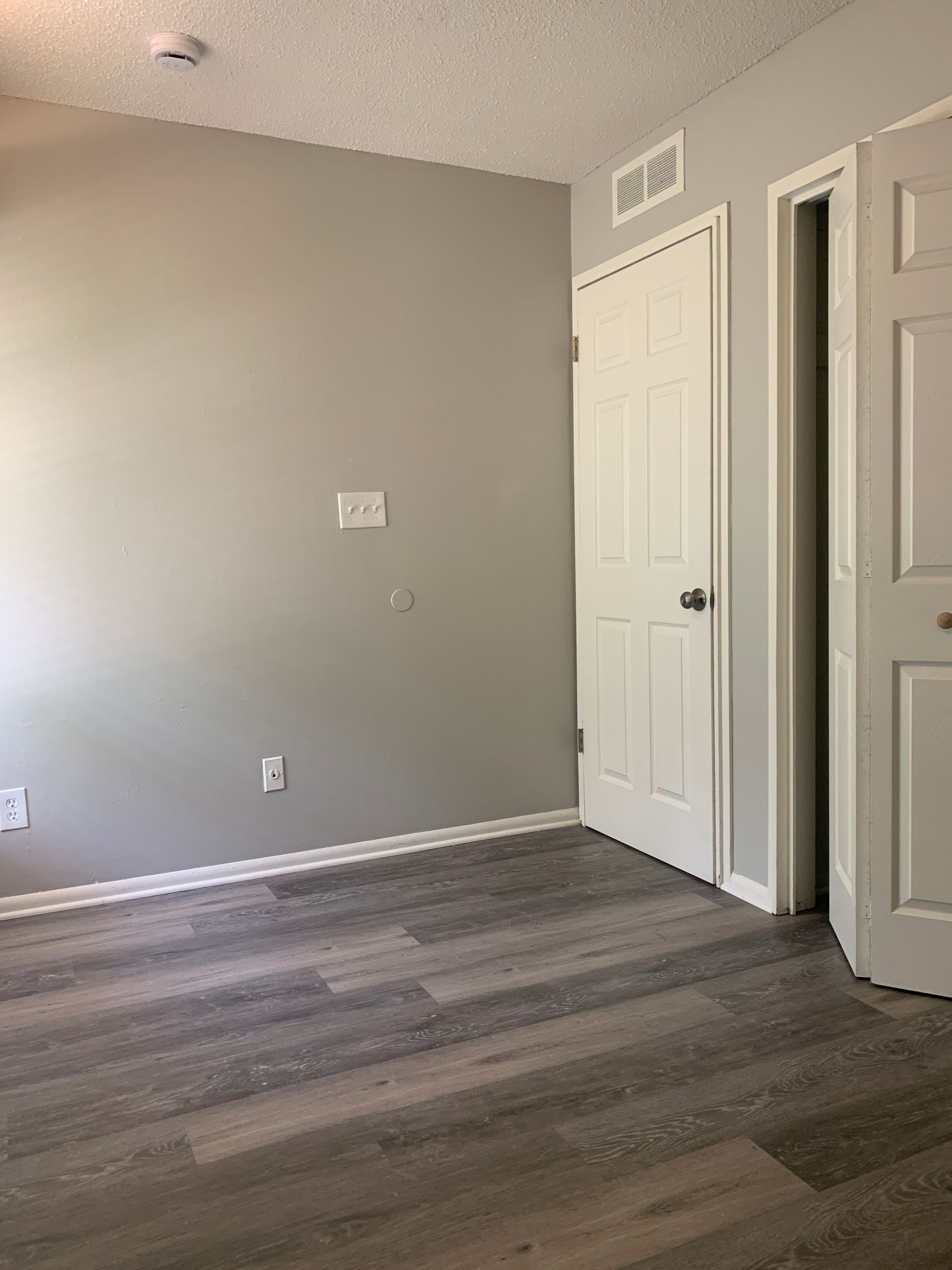 An empty room with gray walls, white trim, and wood-look flooring, a closed door, and an open doorway are visible.