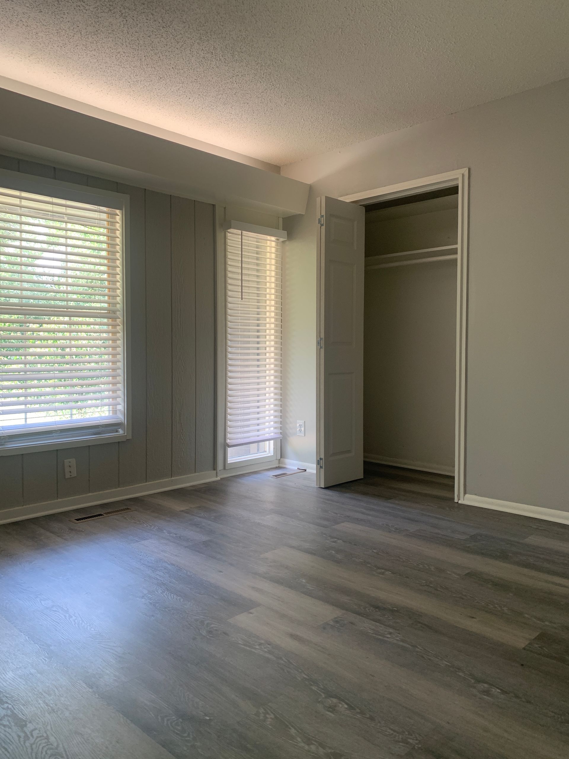 Empty bedroom with gray walls and wood-look flooring. Closet on the right, windows with blinds on the left.