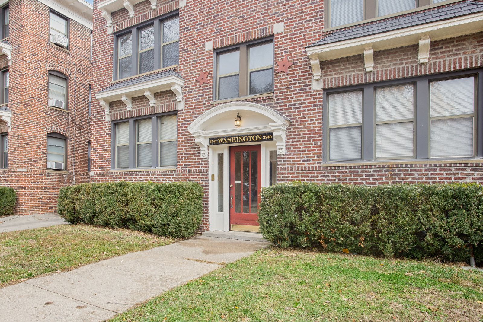 Brick apartment building with green hedges, sidewalk leading to a red door, and windows.