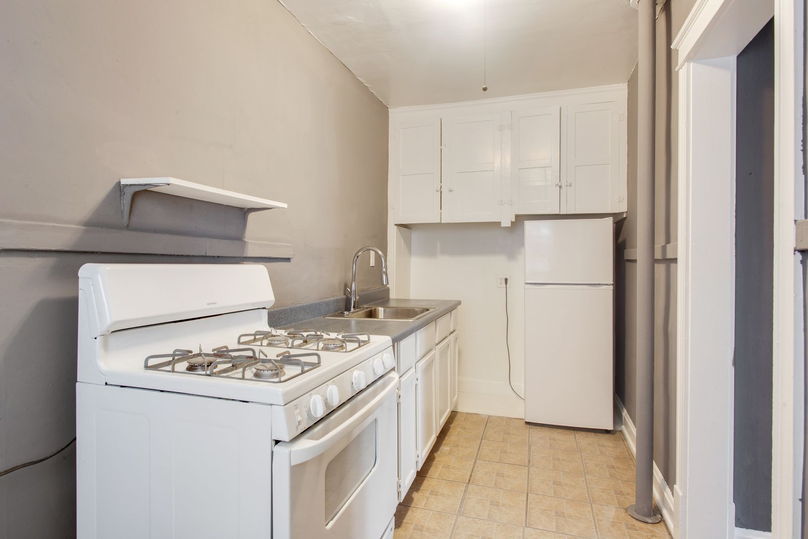 White kitchen with stove, sink, cabinets, and refrigerator, light gray walls, and wooden floor.