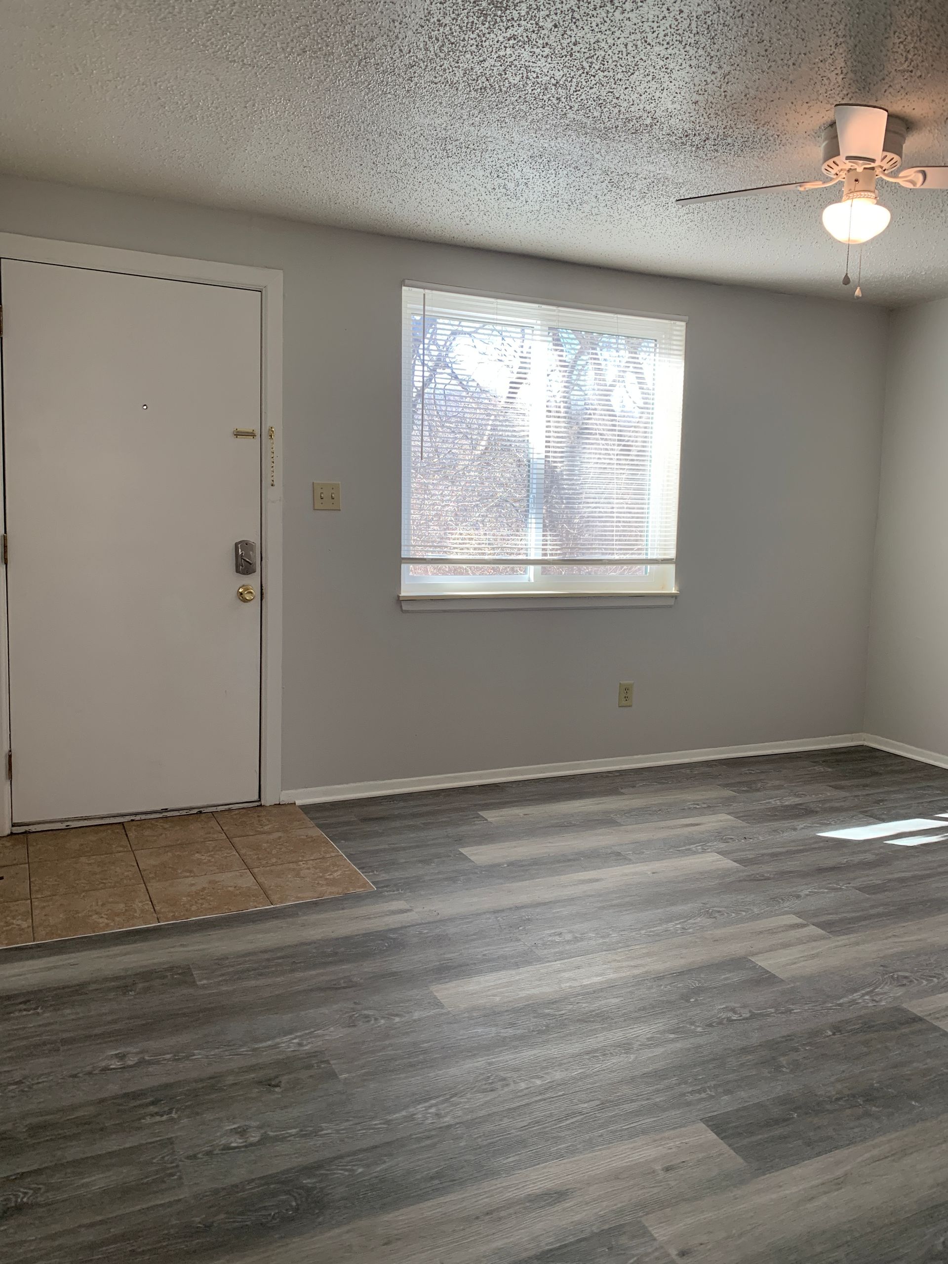 Empty living room with gray walls, wood-look floor, window with blinds, and ceiling fan.