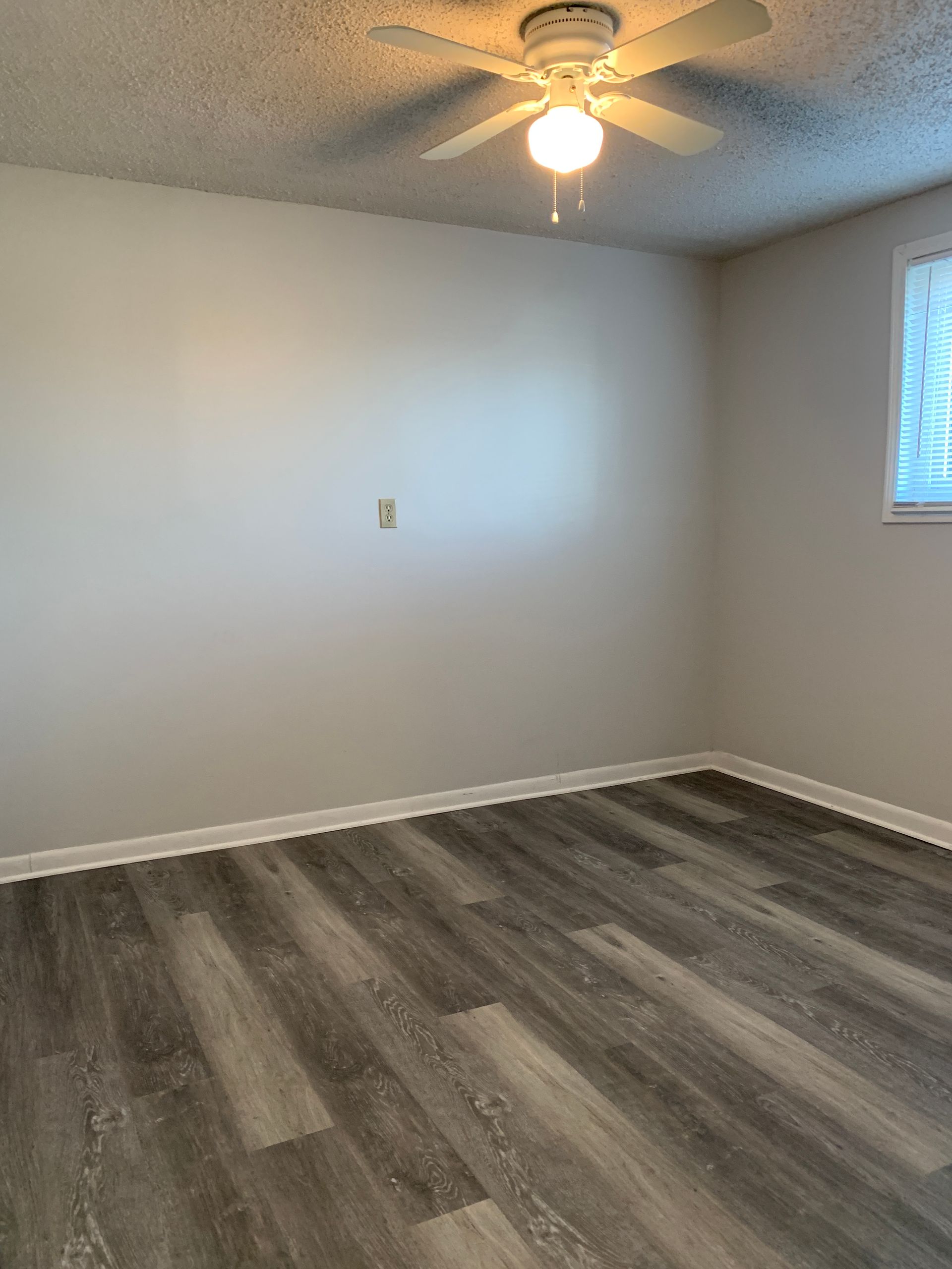 Empty room with gray wood-look flooring, white walls, ceiling fan, and window with blinds.