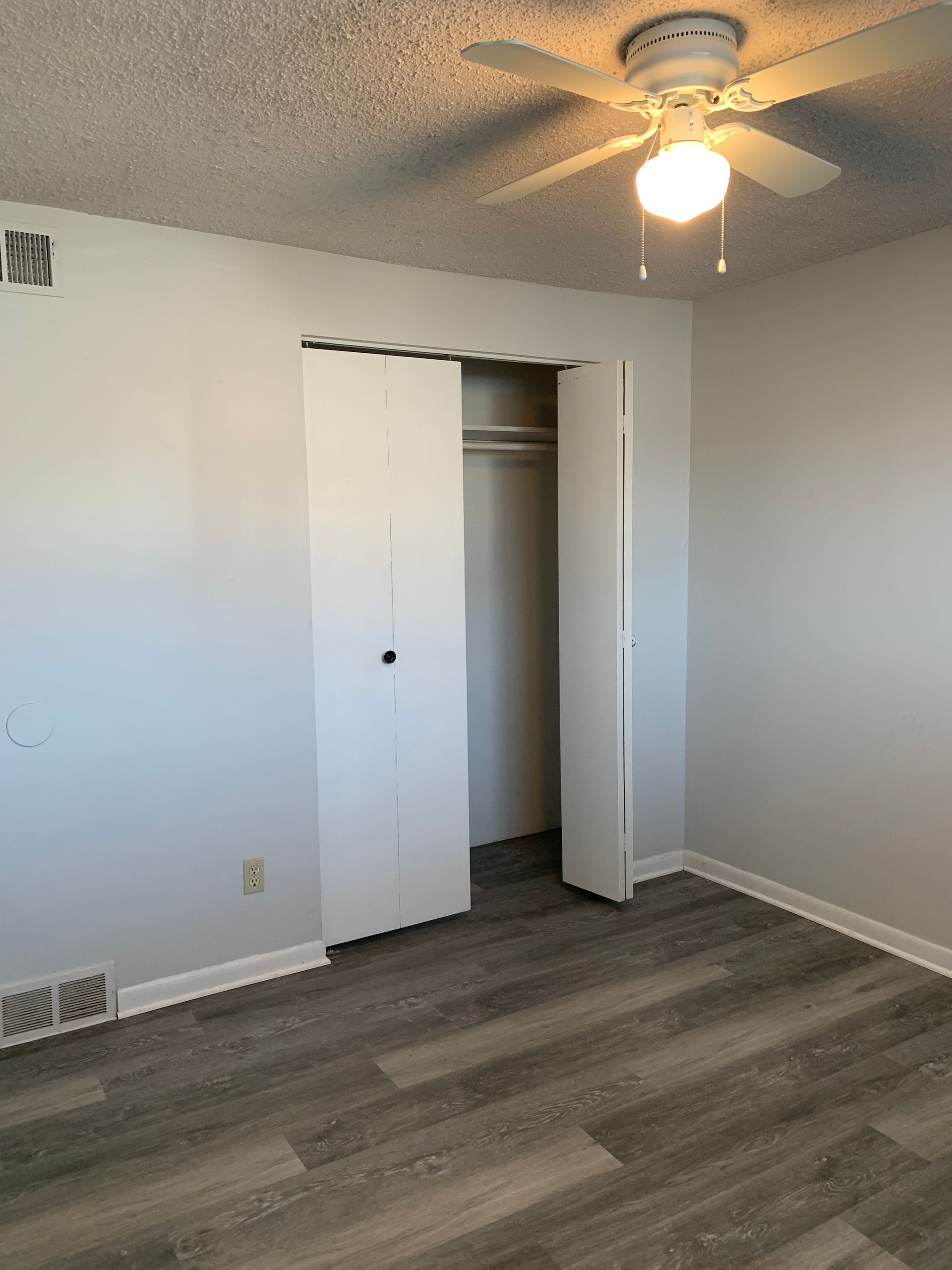 Empty bedroom with gray walls, wood-look floor, and closet with open doors. Ceiling fan with light.
