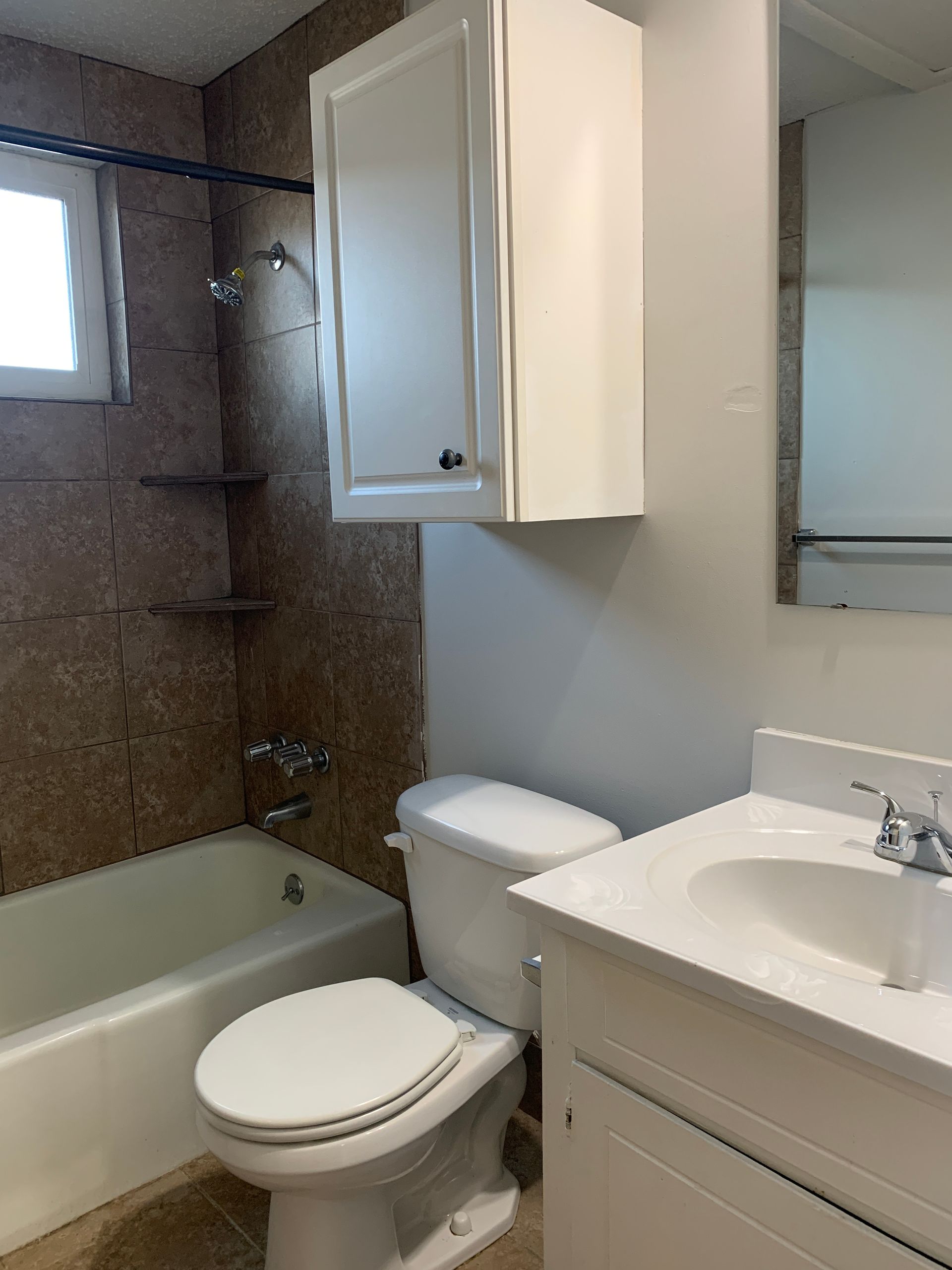 Bathroom with a white toilet, sink, and cabinet. The bathtub has a showerhead and brown tiled walls.