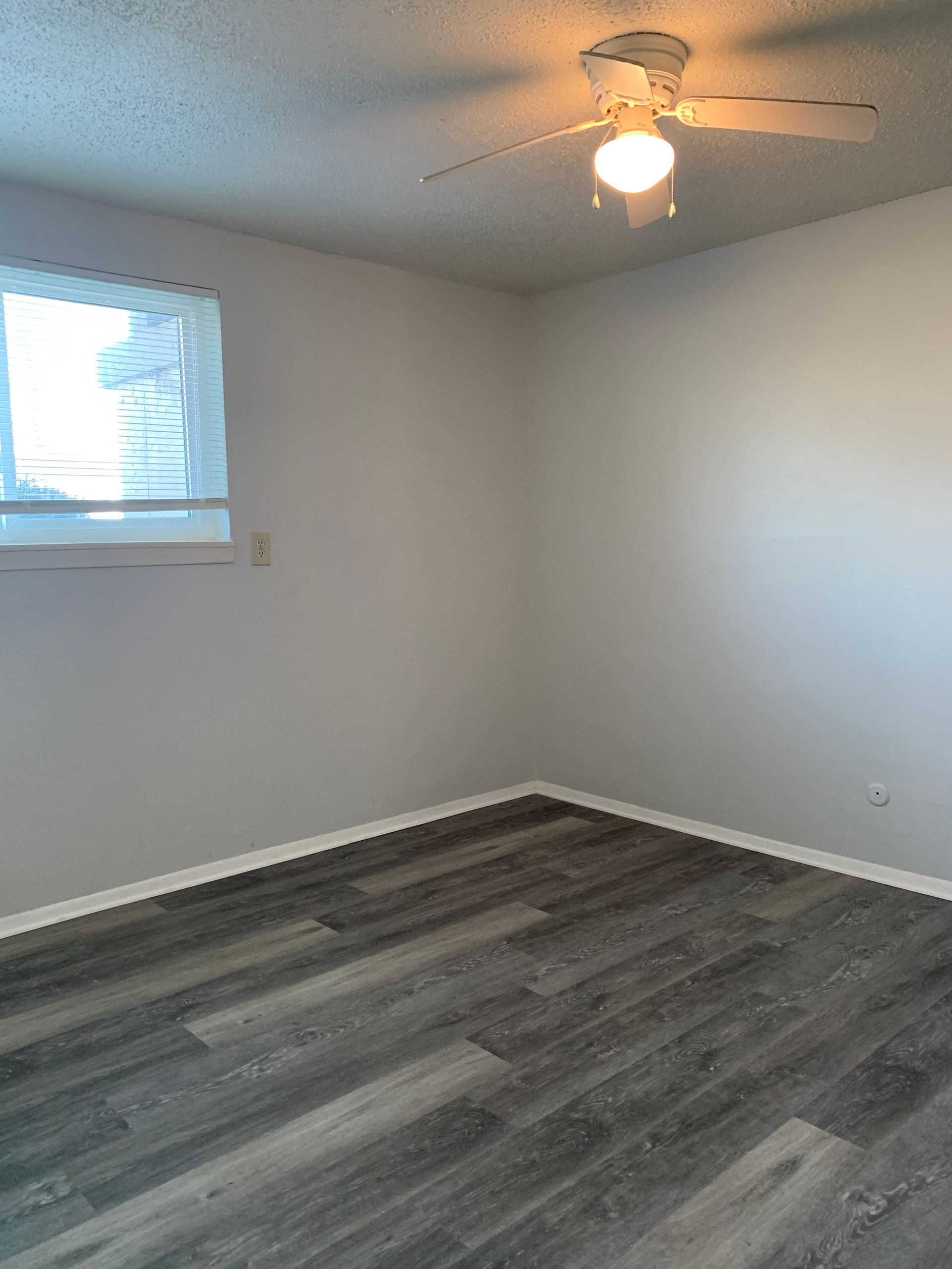 Empty room with gray laminate flooring, white walls, window with blinds, and ceiling fan.