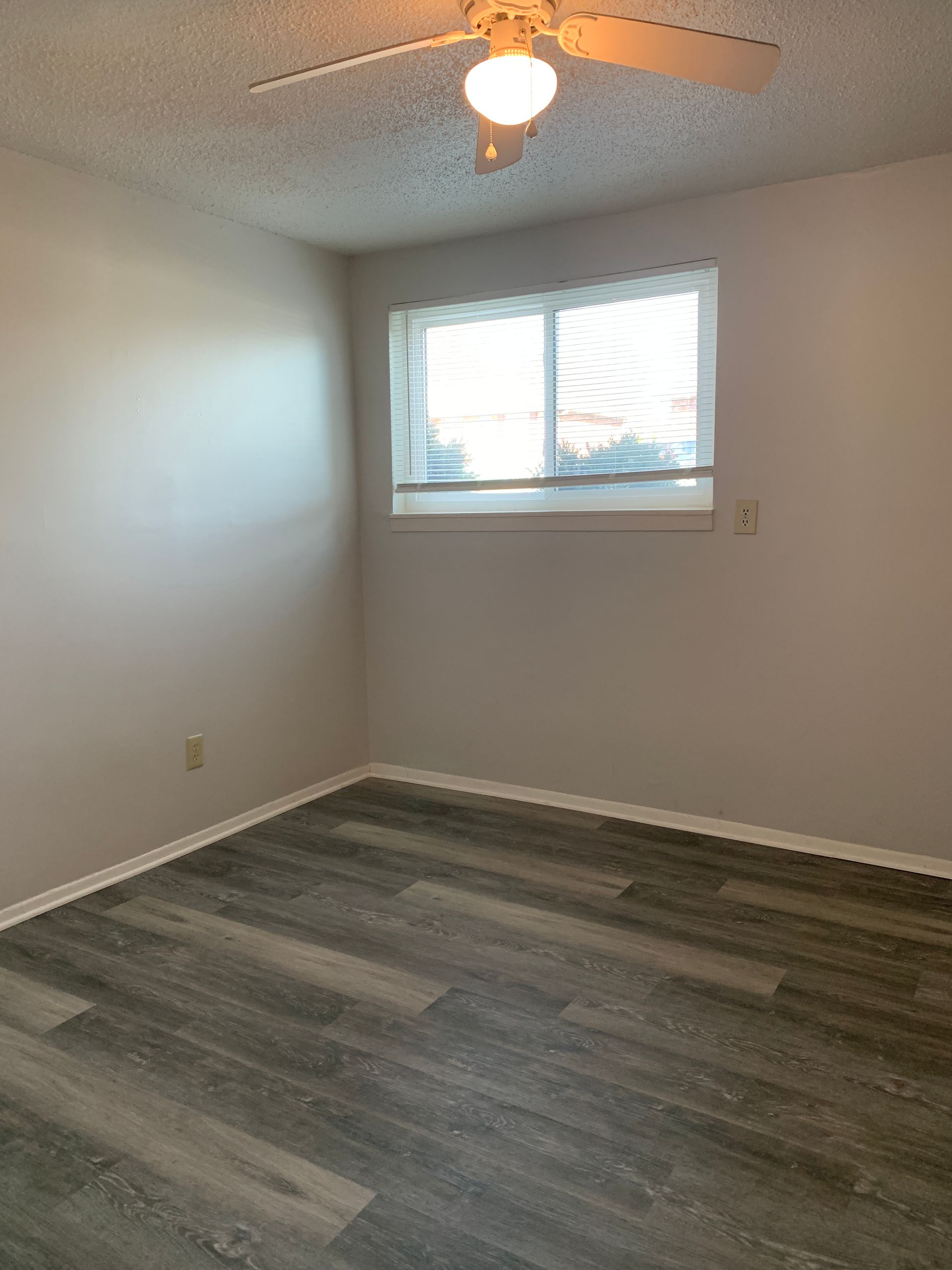 Empty bedroom with gray wood-look flooring, beige walls, and a window with blinds. Ceiling fan present.