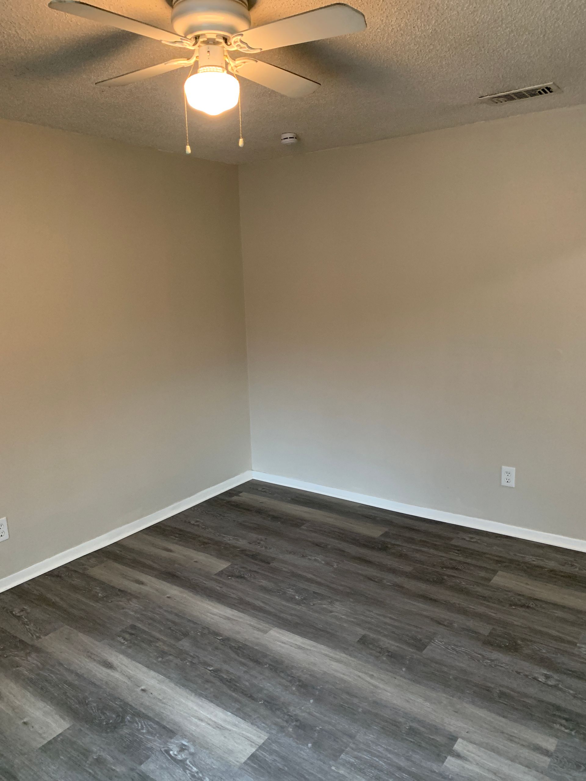 Empty room with gray wood-look flooring, beige walls, and a ceiling fan with a light fixture.