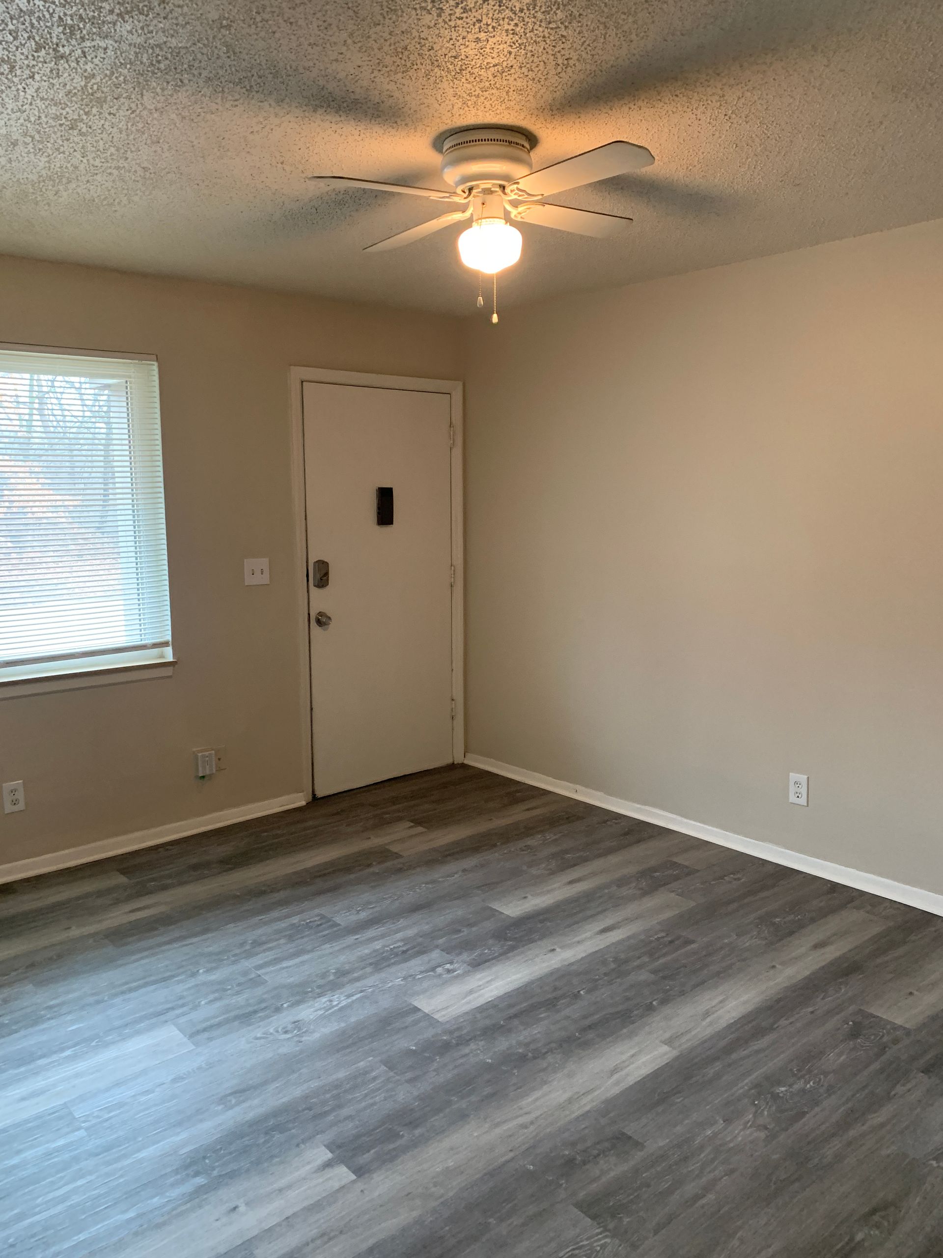 An empty room with gray wood-look flooring, a white door, a window with blinds, and a ceiling fan.