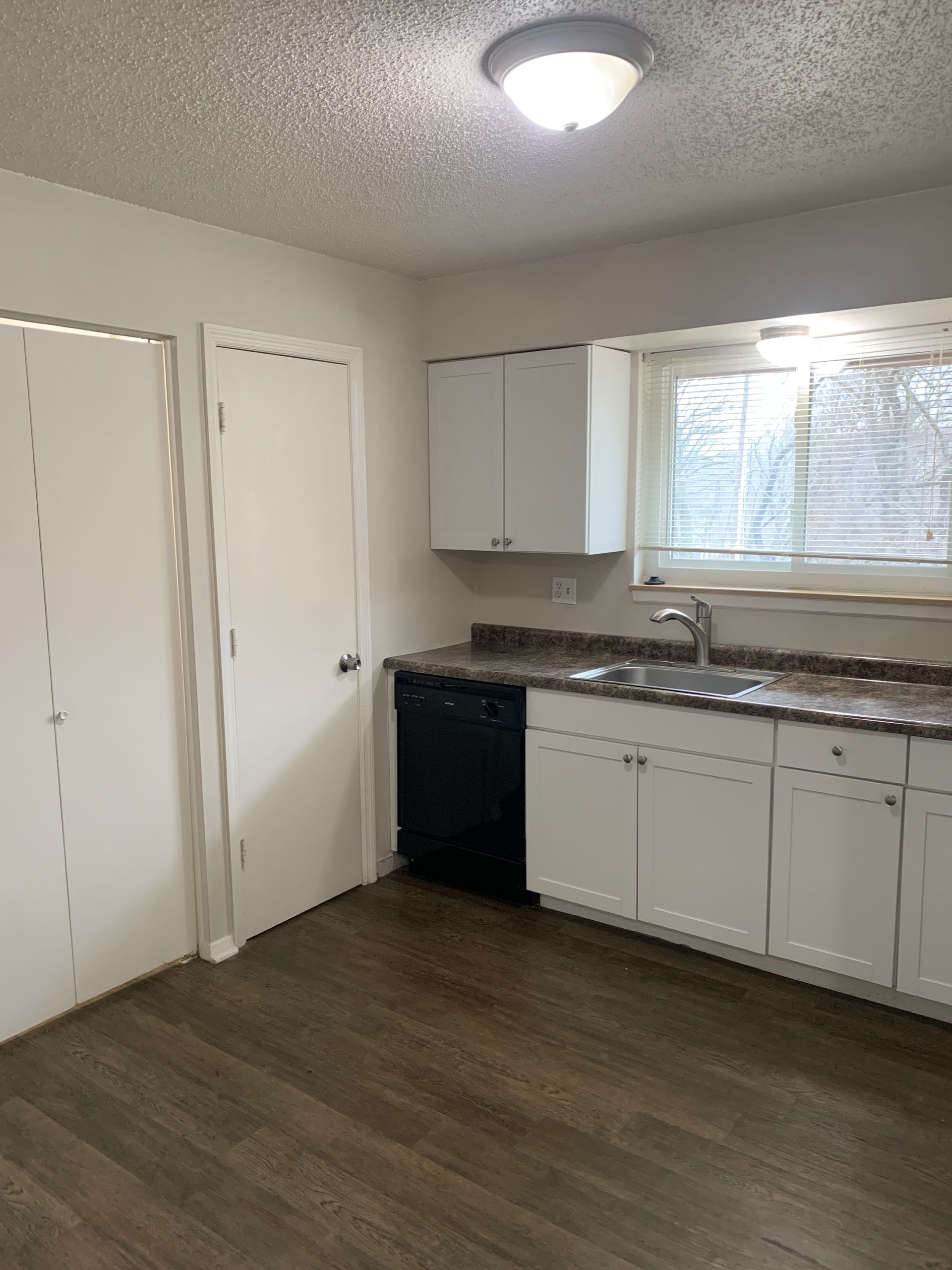 Kitchen with white cabinets, dark countertop, and dark-colored floor. A black dishwasher is present, and a window with blinds.