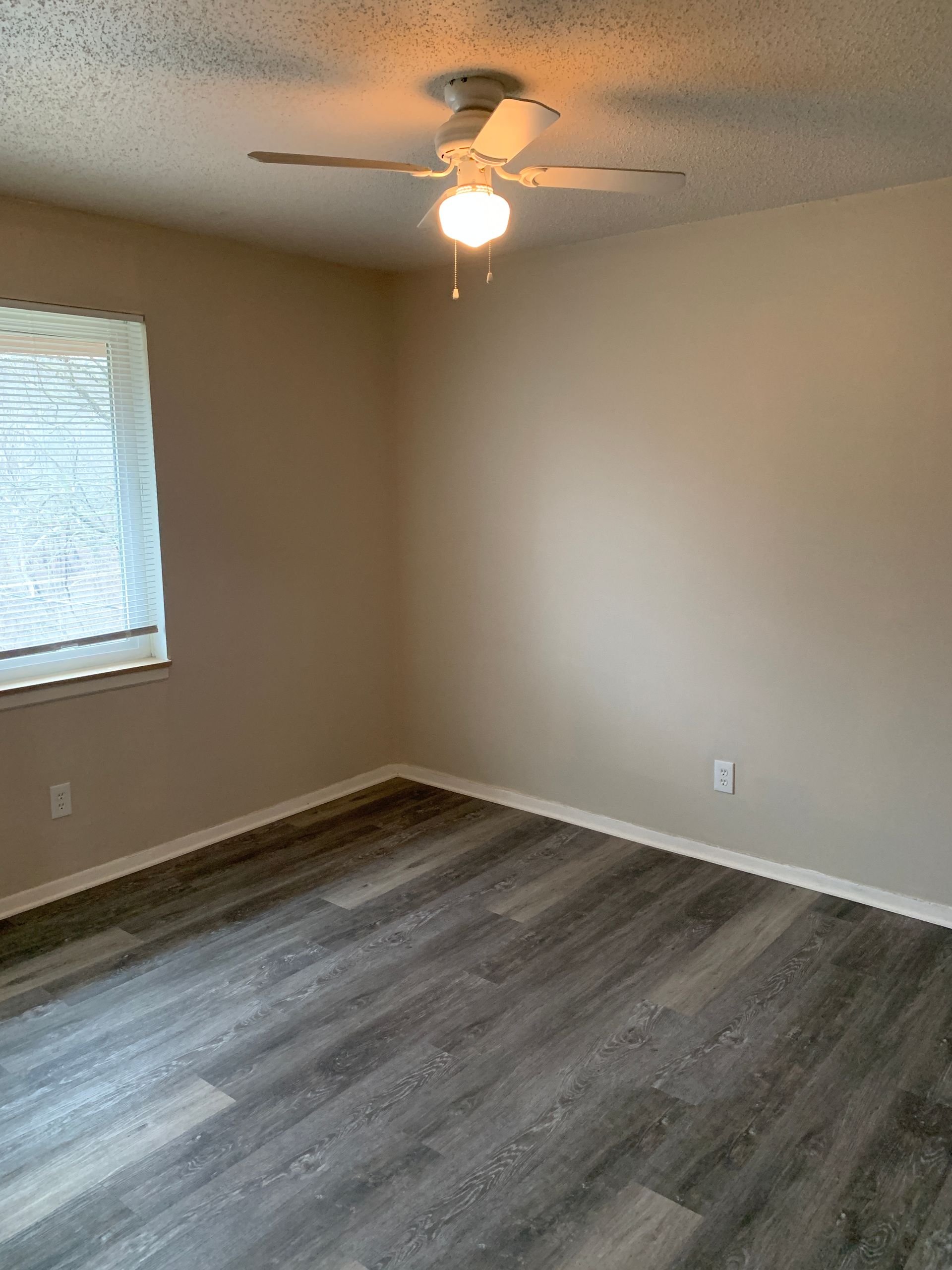 Empty room with gray wood-look flooring, beige walls, a window with blinds, and a ceiling fan.