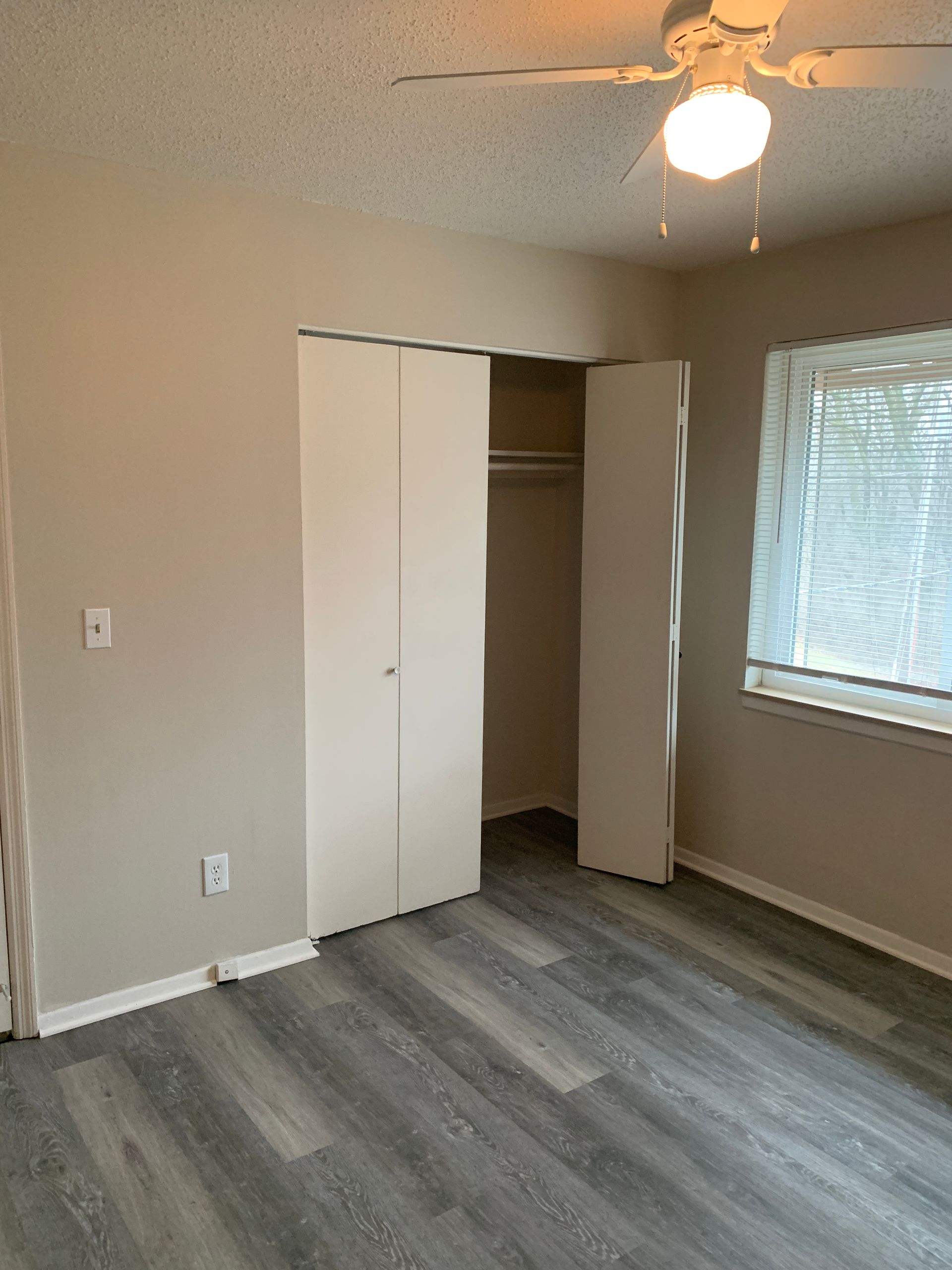 Bedroom with gray wood-look flooring, beige walls, white closet doors, and a window with blinds.