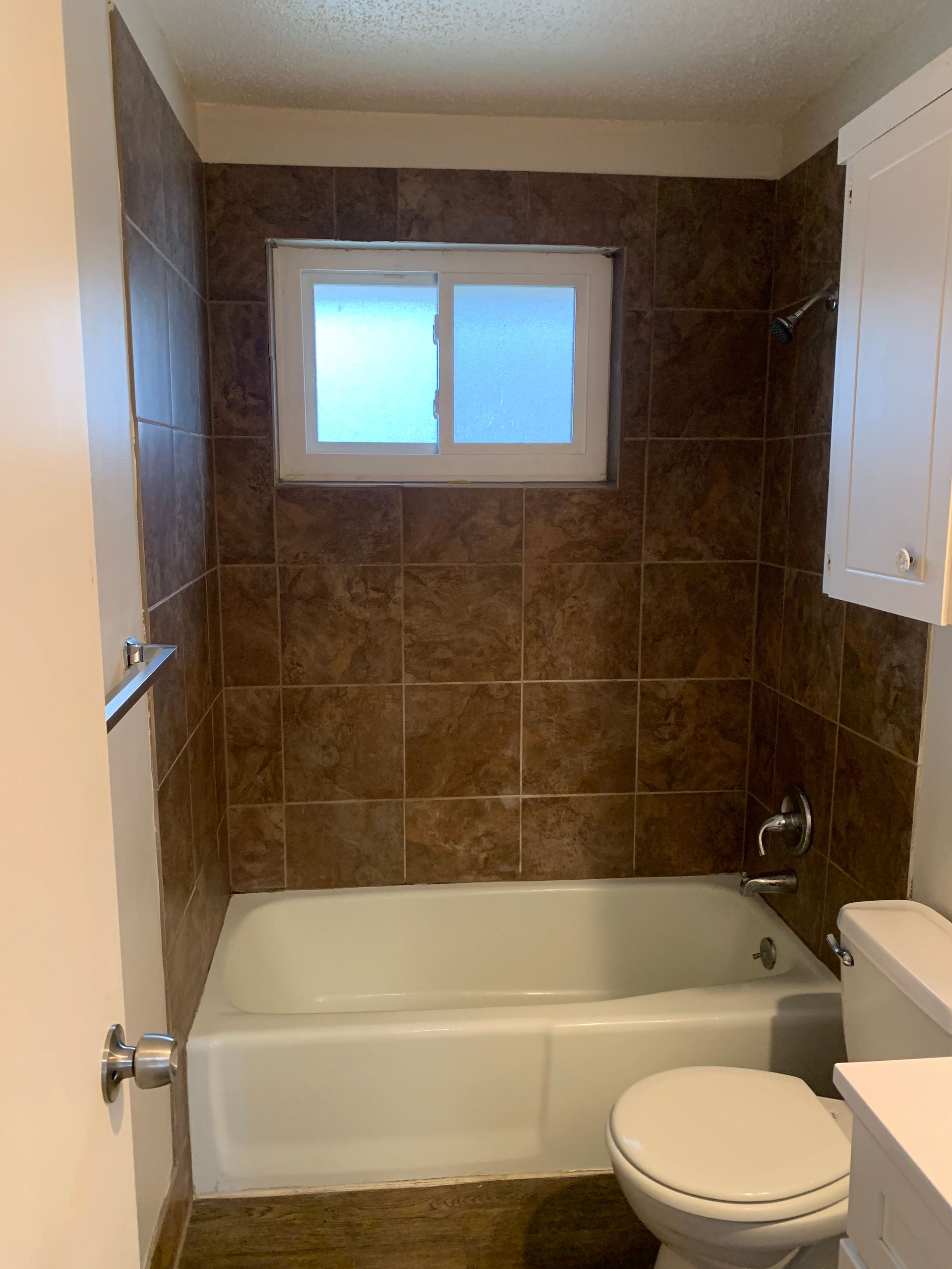Bathroom with a white bathtub, toilet, and cabinet. Brown tile walls, a small window, and a light-colored door.