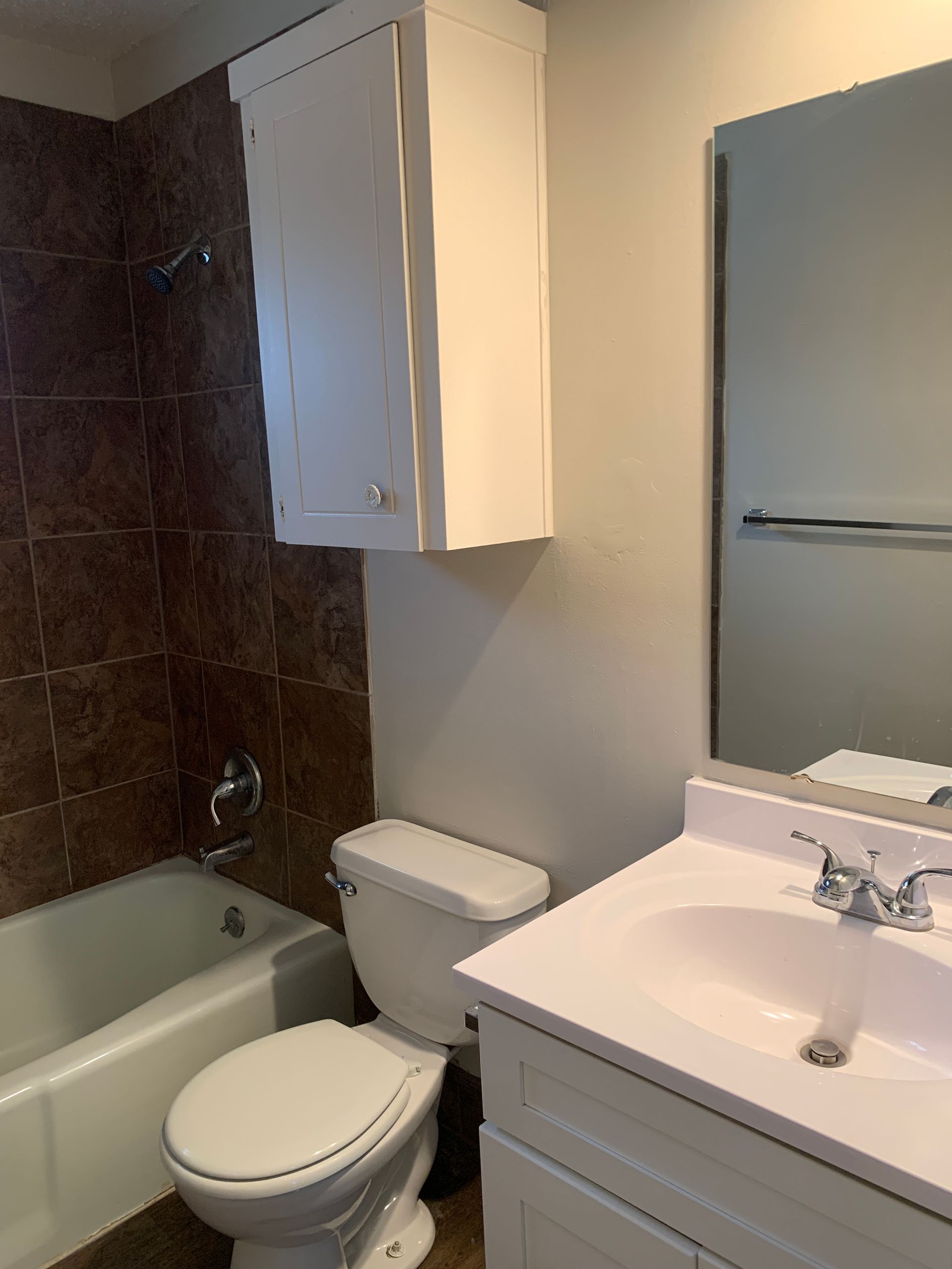 Bathroom with white vanity, toilet, and bathtub. Brown tiled shower wall, white cabinet, and large mirror.
