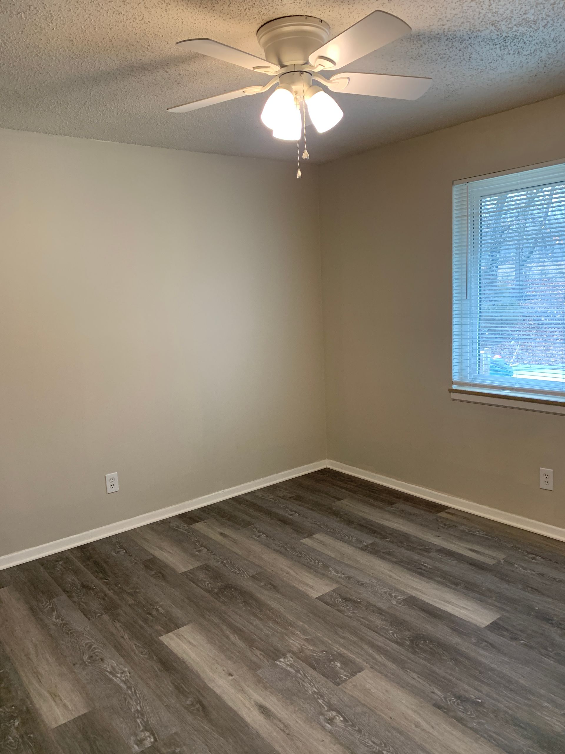 Empty bedroom with gray wood-look flooring, beige walls, a window, and a ceiling fan.