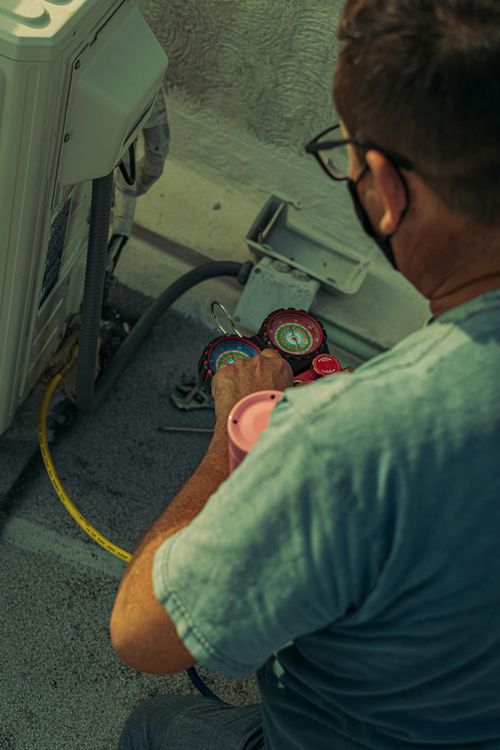 Person in a mask servicing AC unit with gauges. Gray shirt, outdoors, sunlight.