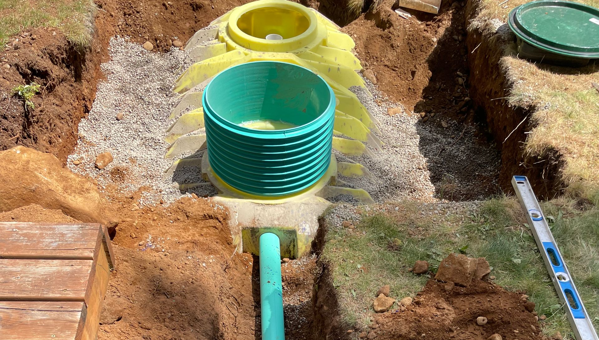 Excavated area with yellow and green septic system components, connected by a green pipe, in a yard.
