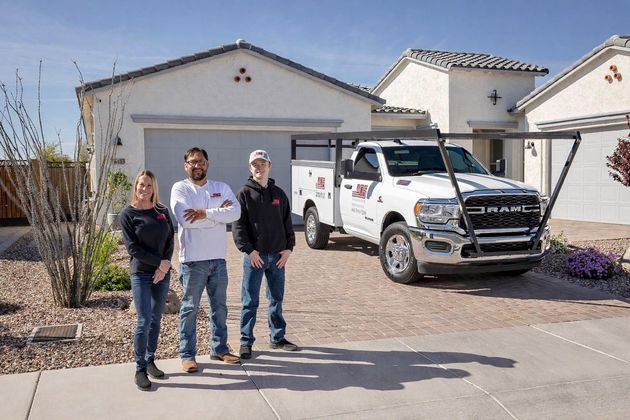 Three people stand smiling in front of a white utility truck parked at a suburban house on a sunny day.