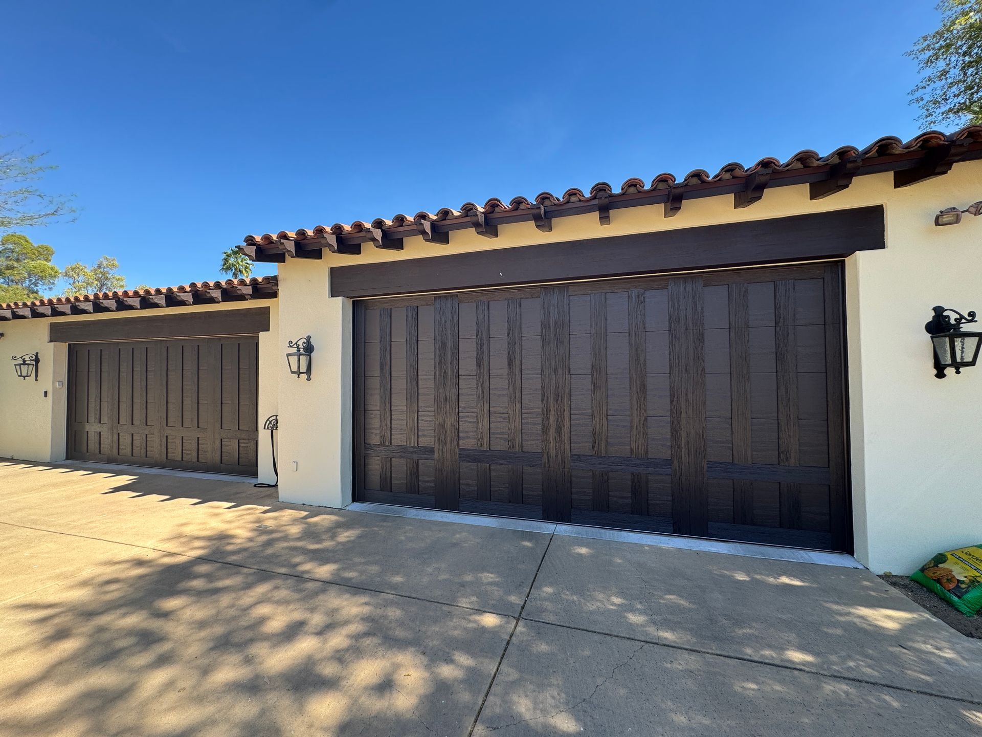 A two-car garage with dark wood panel doors, cream-colored stucco walls, and a Spanish-style tile roof under a blue sky.