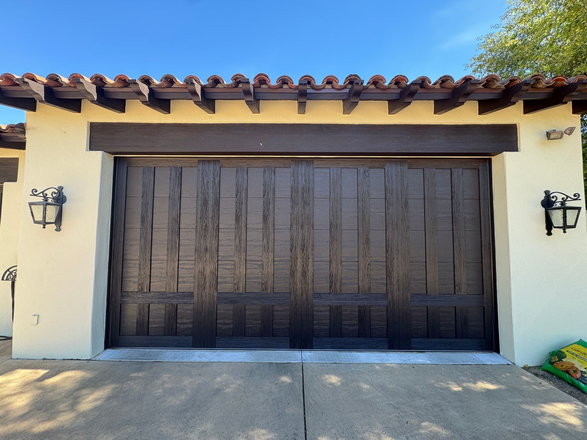 Dark wood garage door with vertical paneling, set in a cream stucco wall with exterior lanterns and a tiled roof edge.