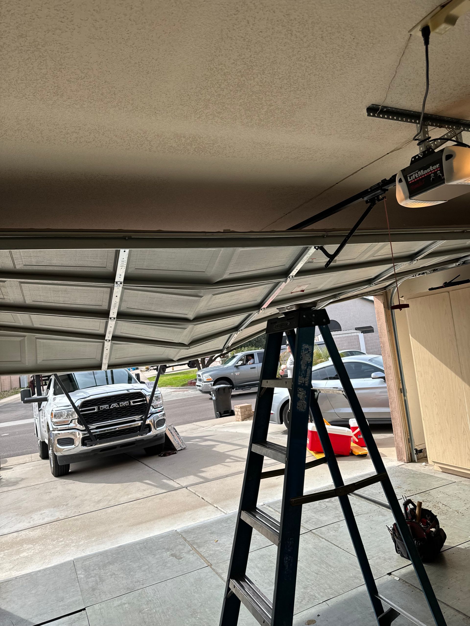 A residential garage door has buckled and bent inward while halfway open, with a ladder and driveway visible in the scene.