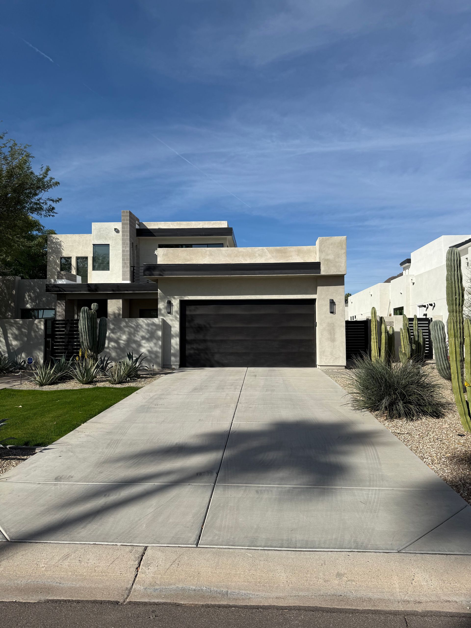 A modern two-story home with a dark garage door, concrete driveway, and desert landscaping under a bright blue sky.