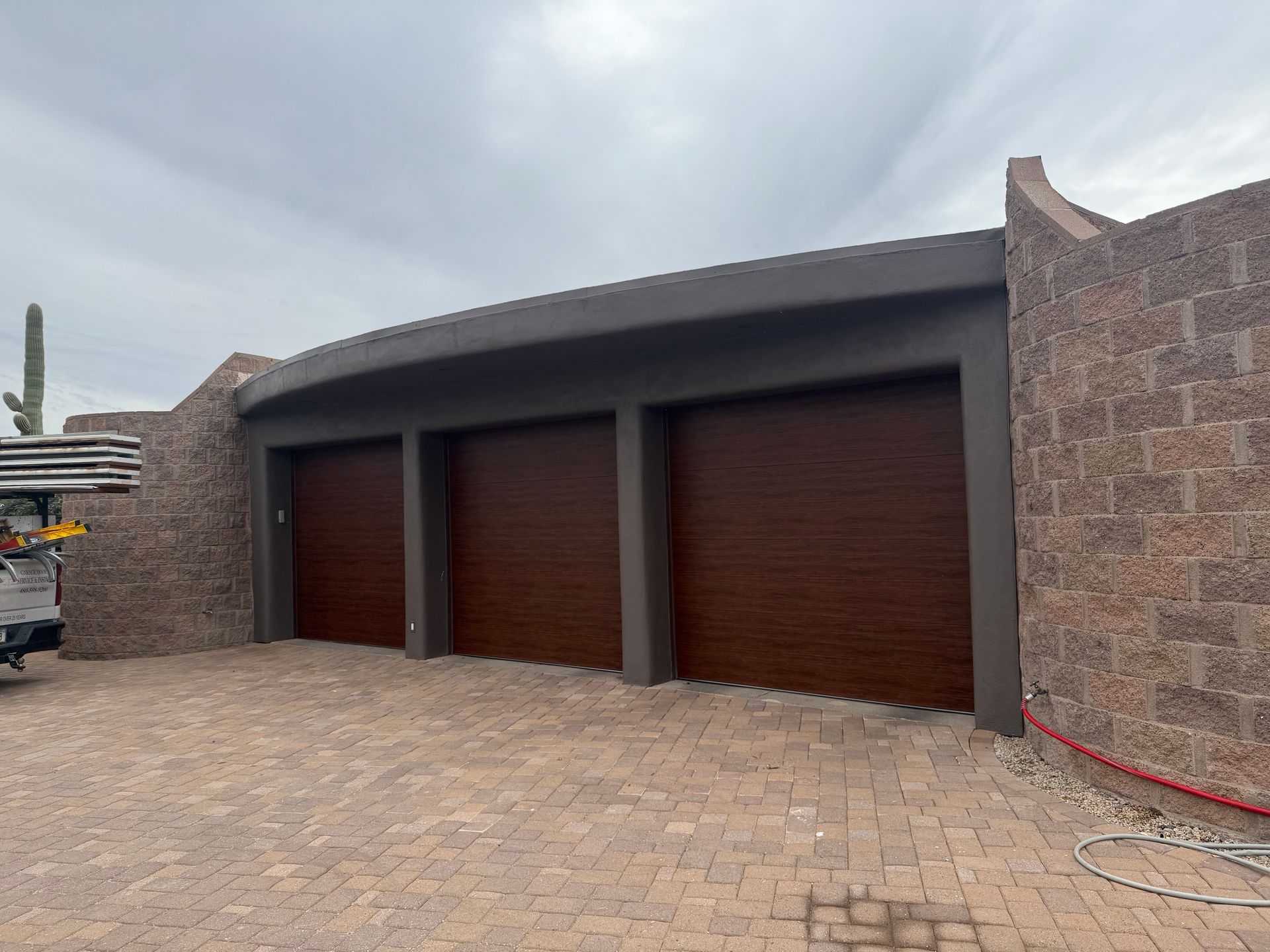 Three modern, brown wood-grain garage doors set into a dark gray stucco structure with stone block side walls.