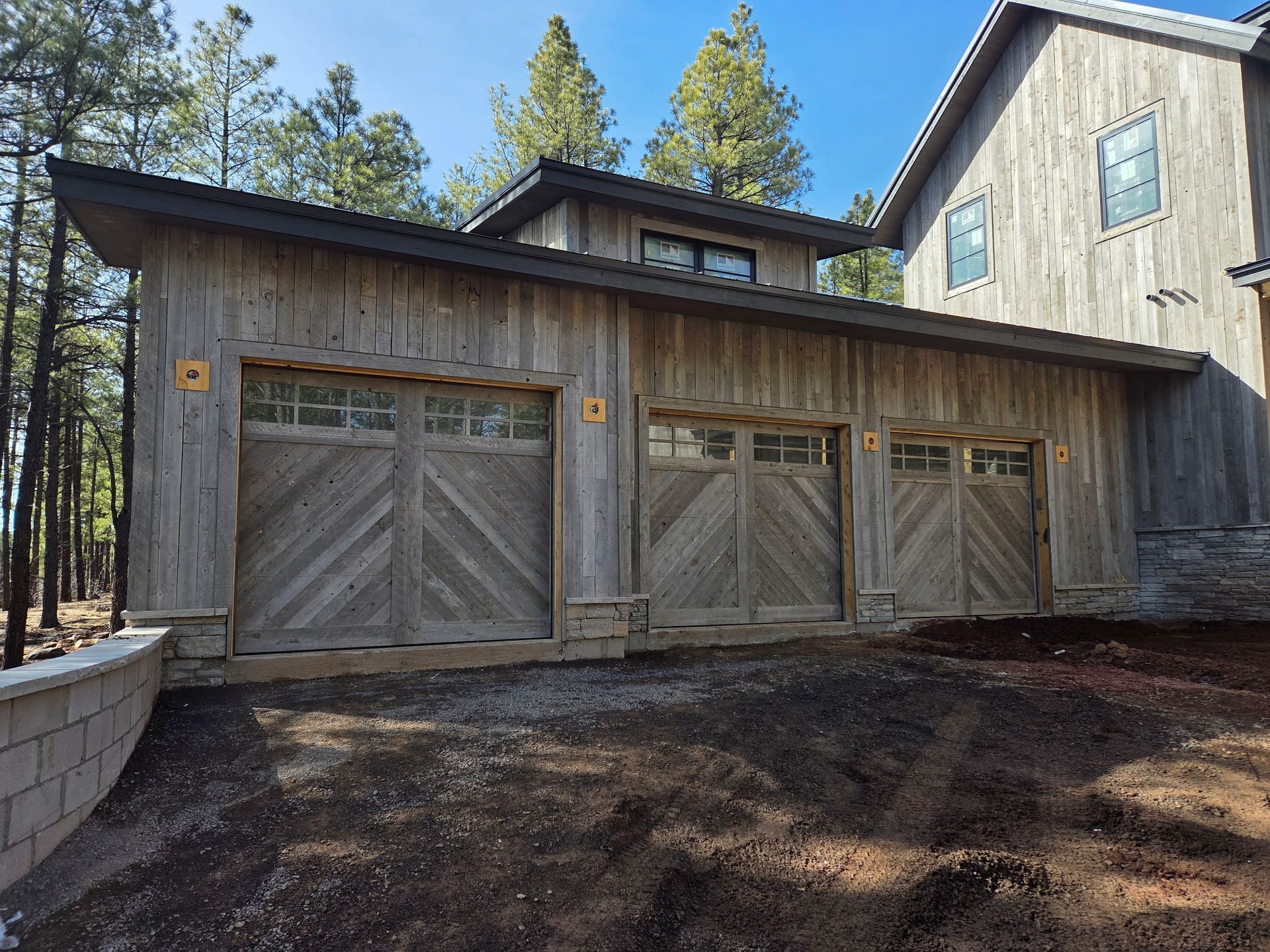 A three-car garage with weathered wood siding, diagonal board doors, and a stone foundation, set in a forested area.