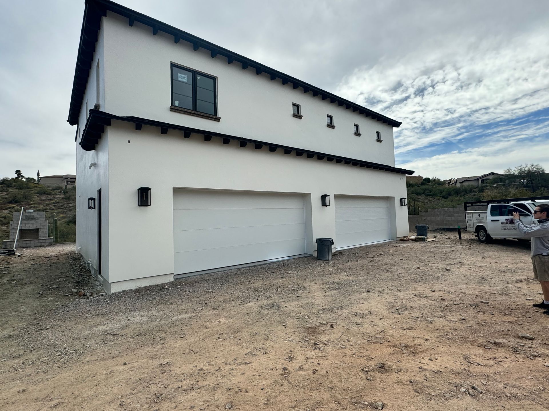 A two-story white building with black trim and two garage doors, situated on a dirt lot under a partly cloudy sky.