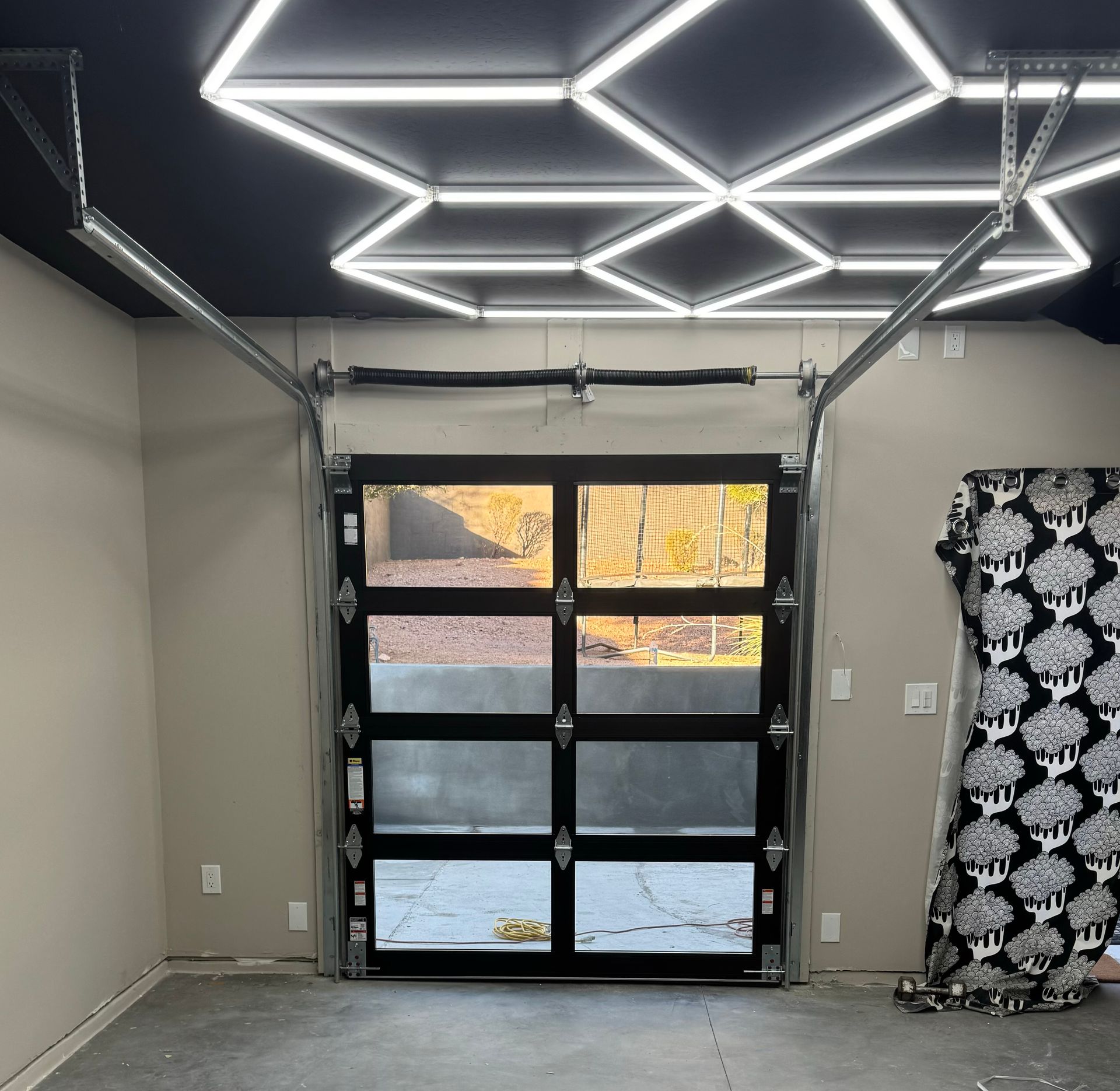 A garage interior features a geometric LED ceiling light grid above a black-framed, multi-pane glass garage door.