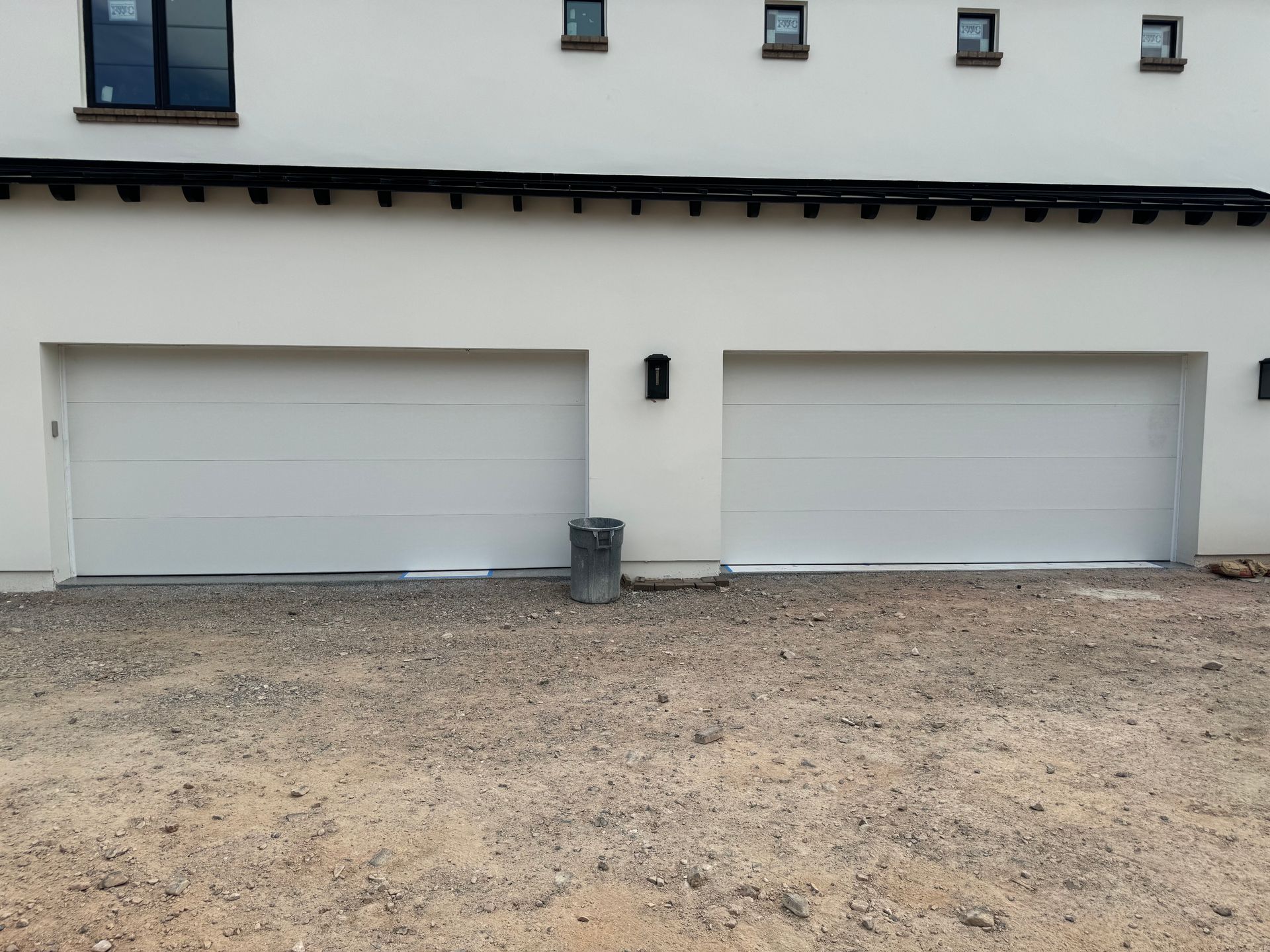 Modern white home with two large garage doors, black light fixtures, and windows above, set over a gravel driveway.