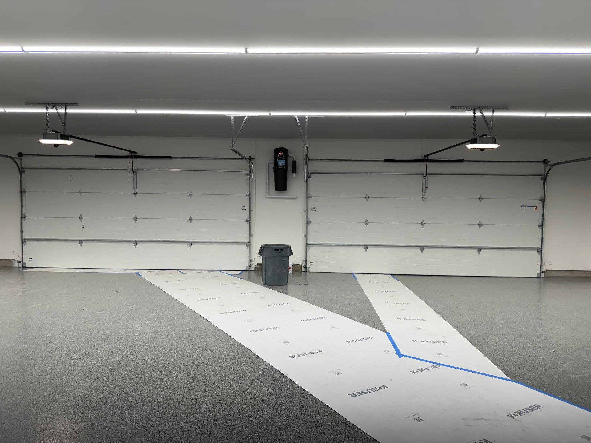 A garage interior with two closed white doors, a central trash bin, and protective white floor paper strips.