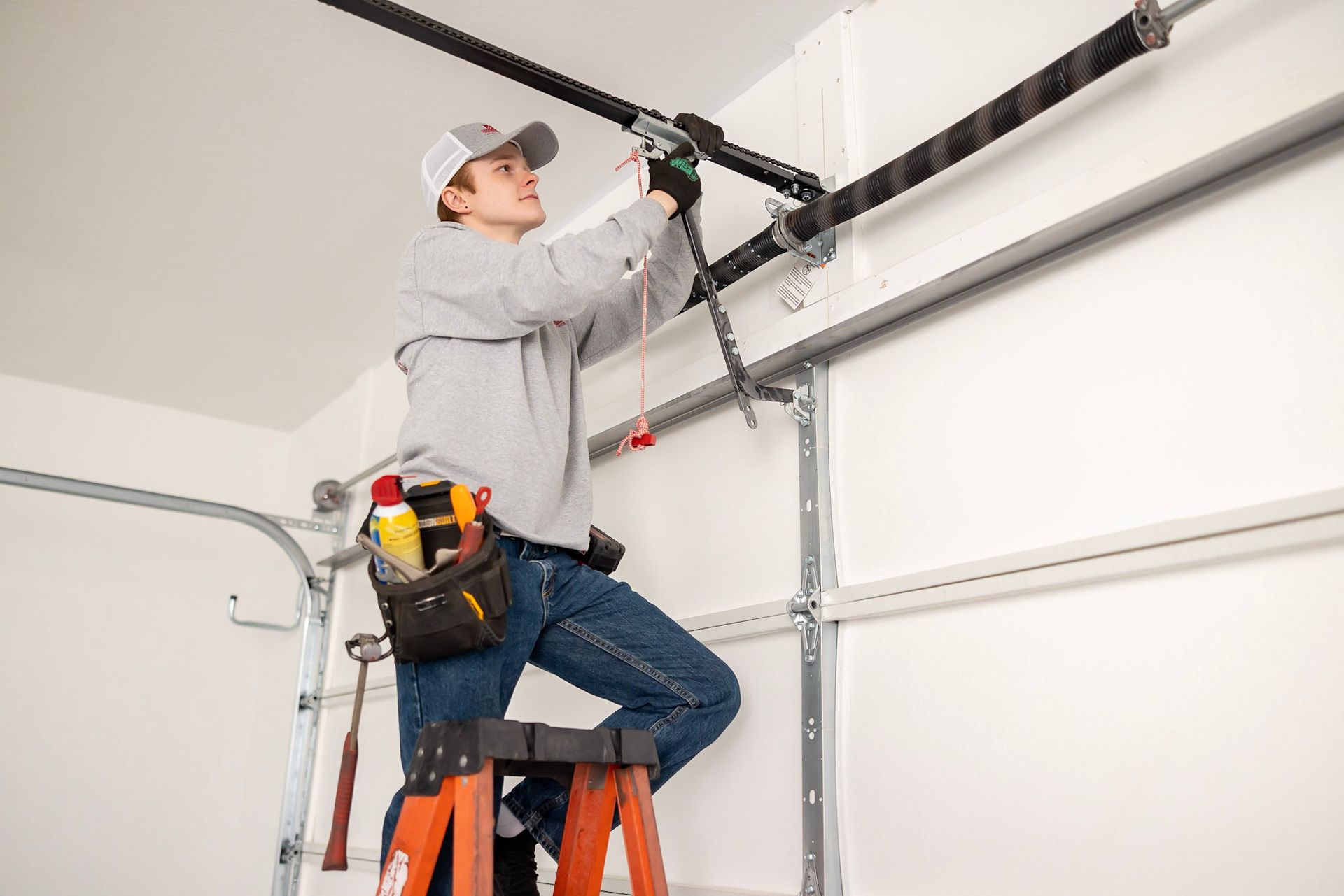 A person wearing a tool belt stands on a ladder, using tools to repair a residential garage door torsion spring system.