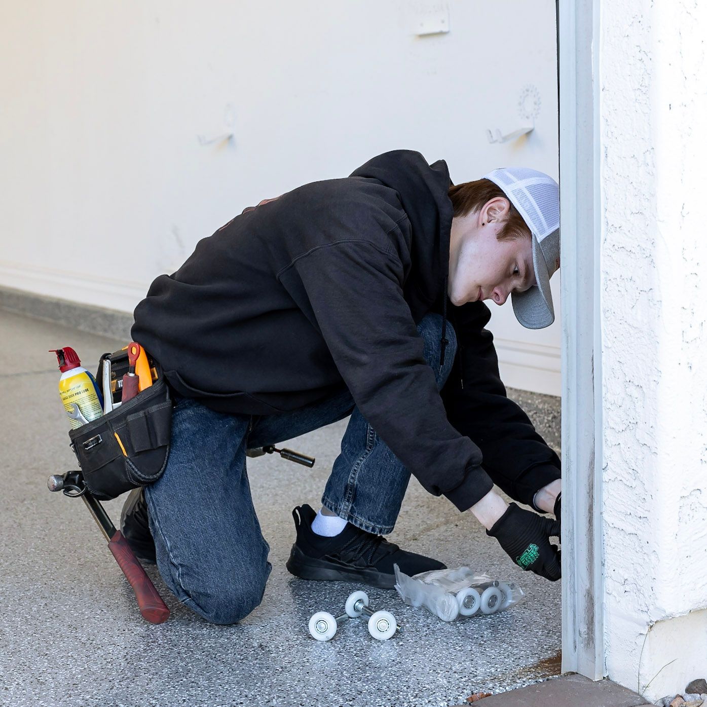 A person in a black hoodie kneels on a garage floor, working on installing garage door rollers into a track.