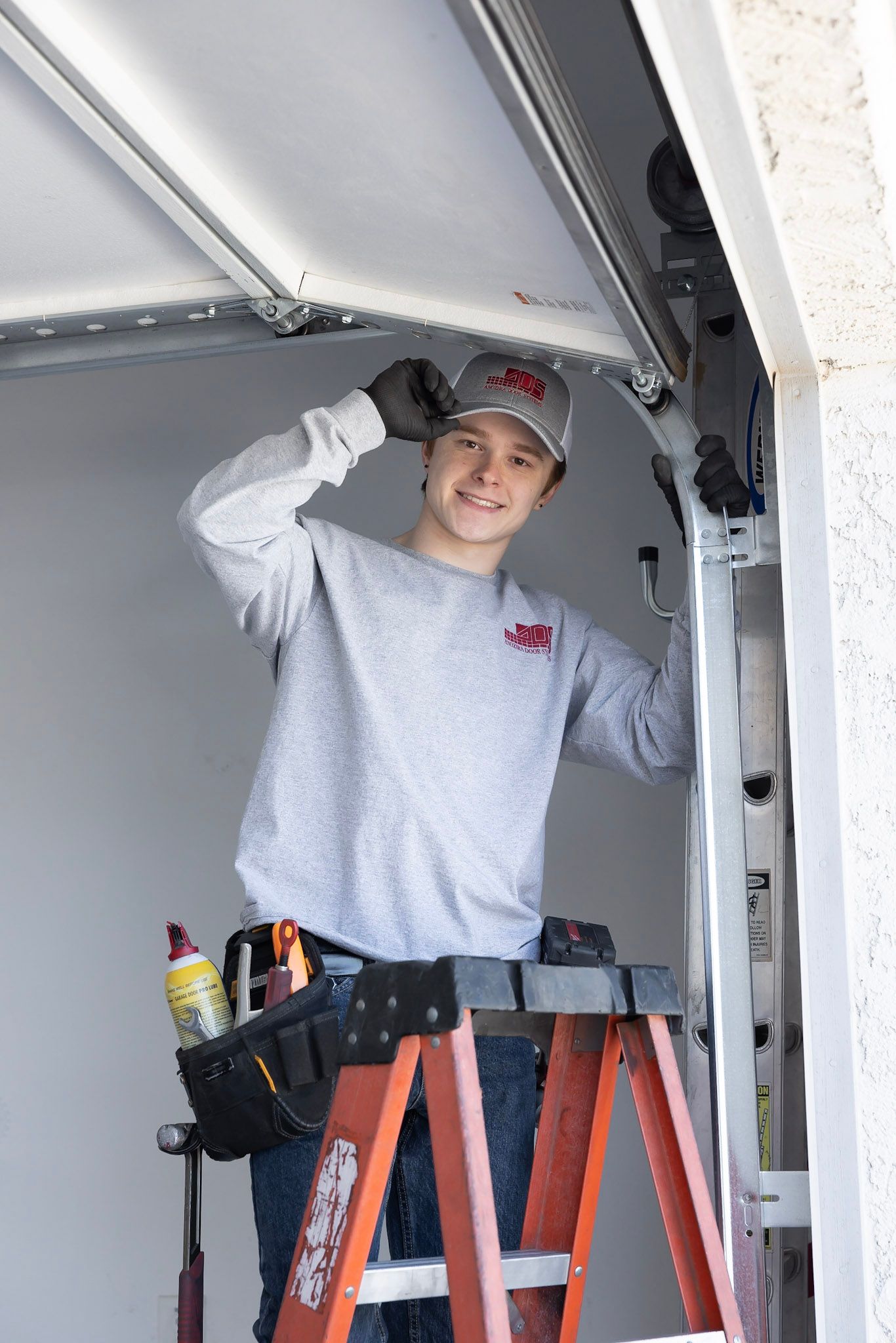 A person wearing a grey sweater, cap, and tool belt stands on a step ladder, working on a garage door track.