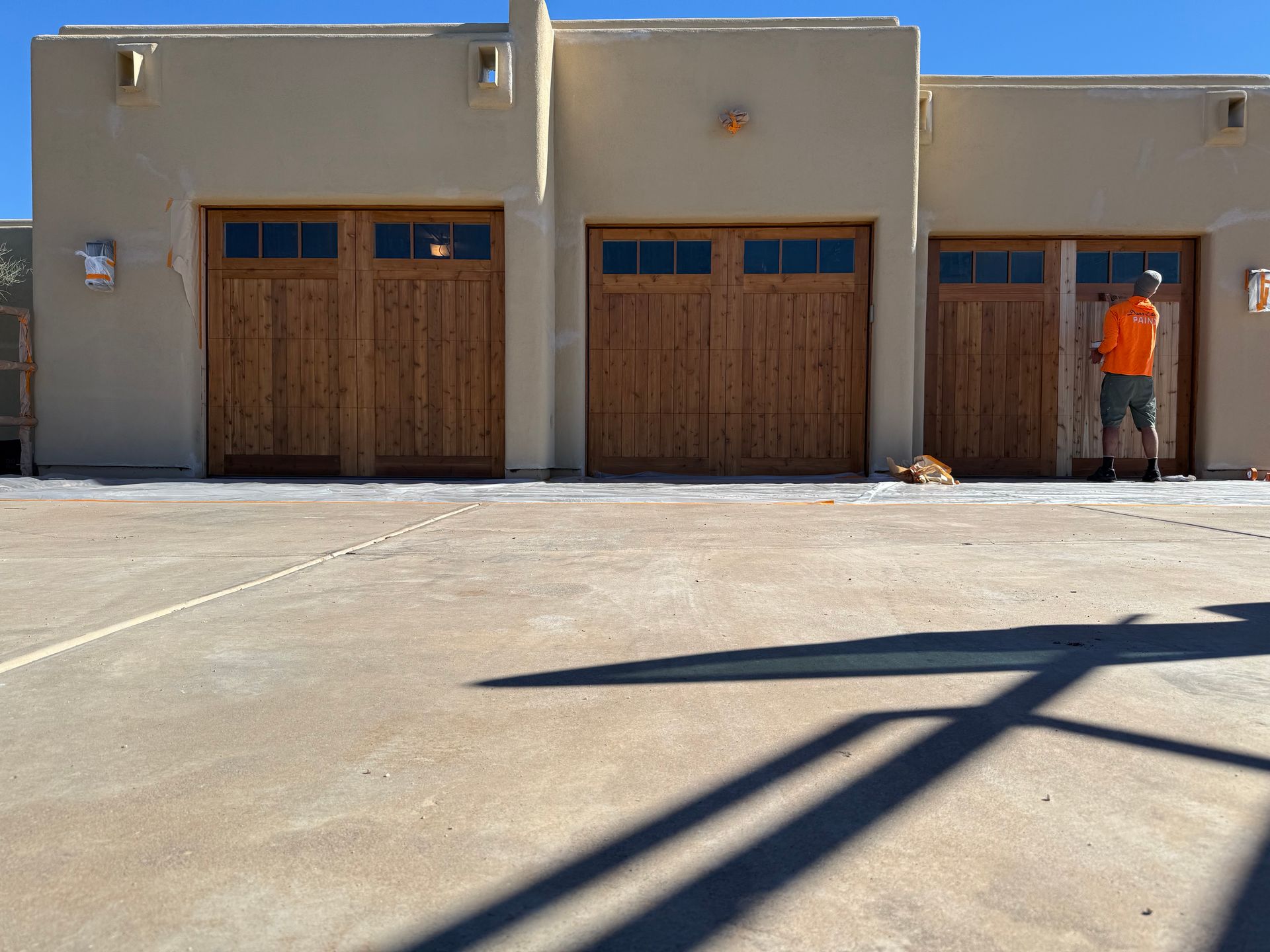 A person wearing an orange shirt stands by the right garage door of a stucco home with three wooden garage doors.