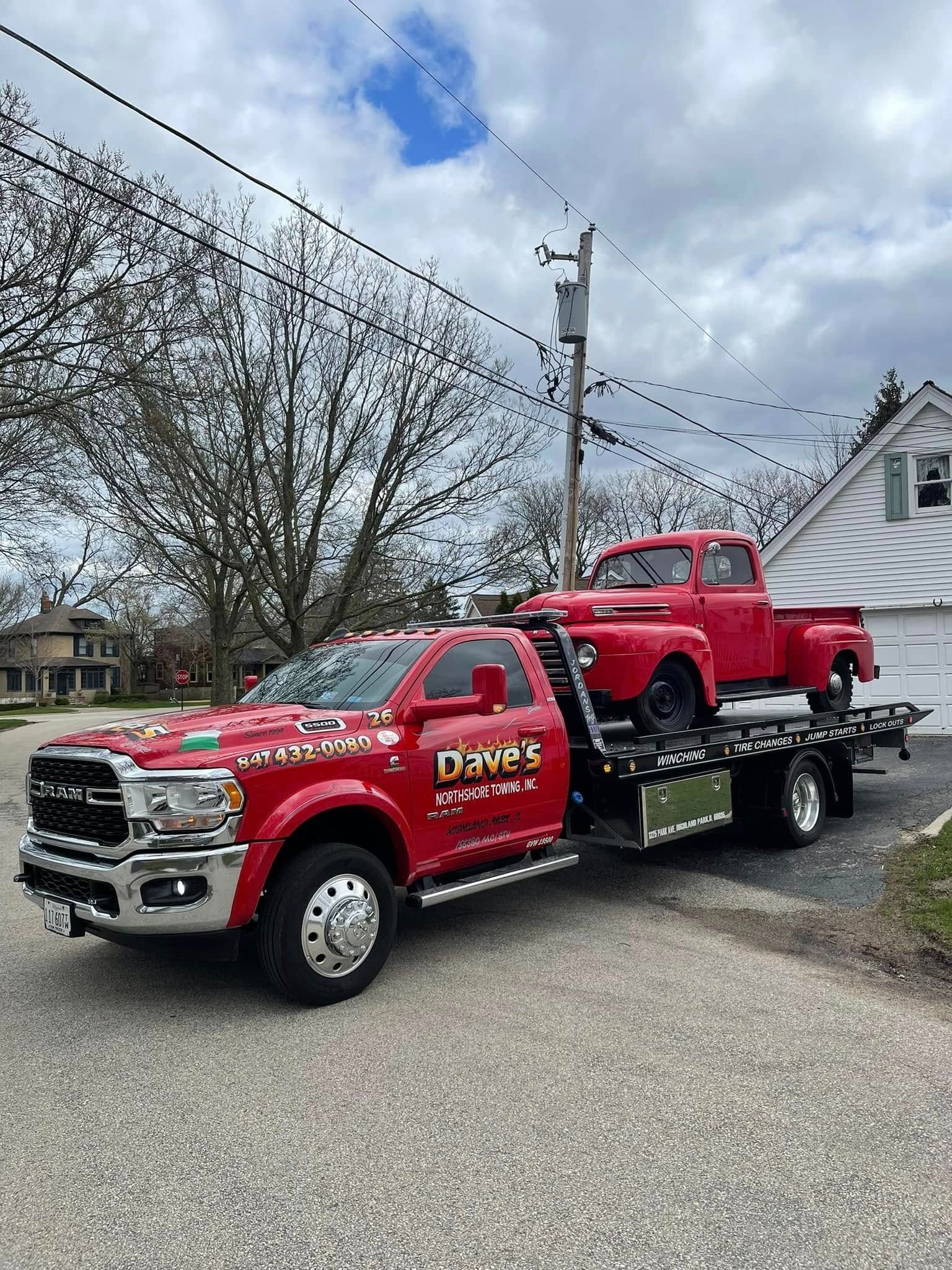 A red tow truck is towing a red truck in front of a house.