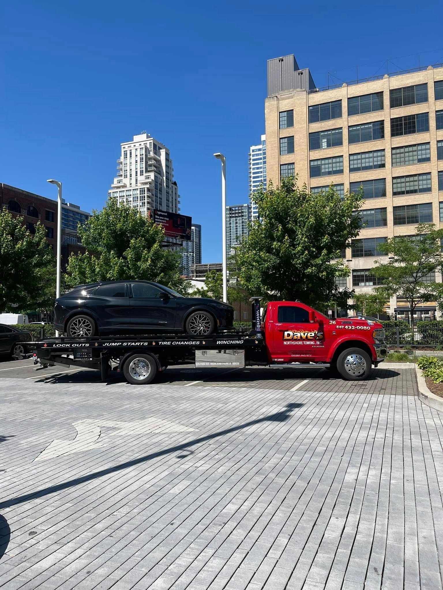 A red tow truck is towing a black car in a parking lot.
