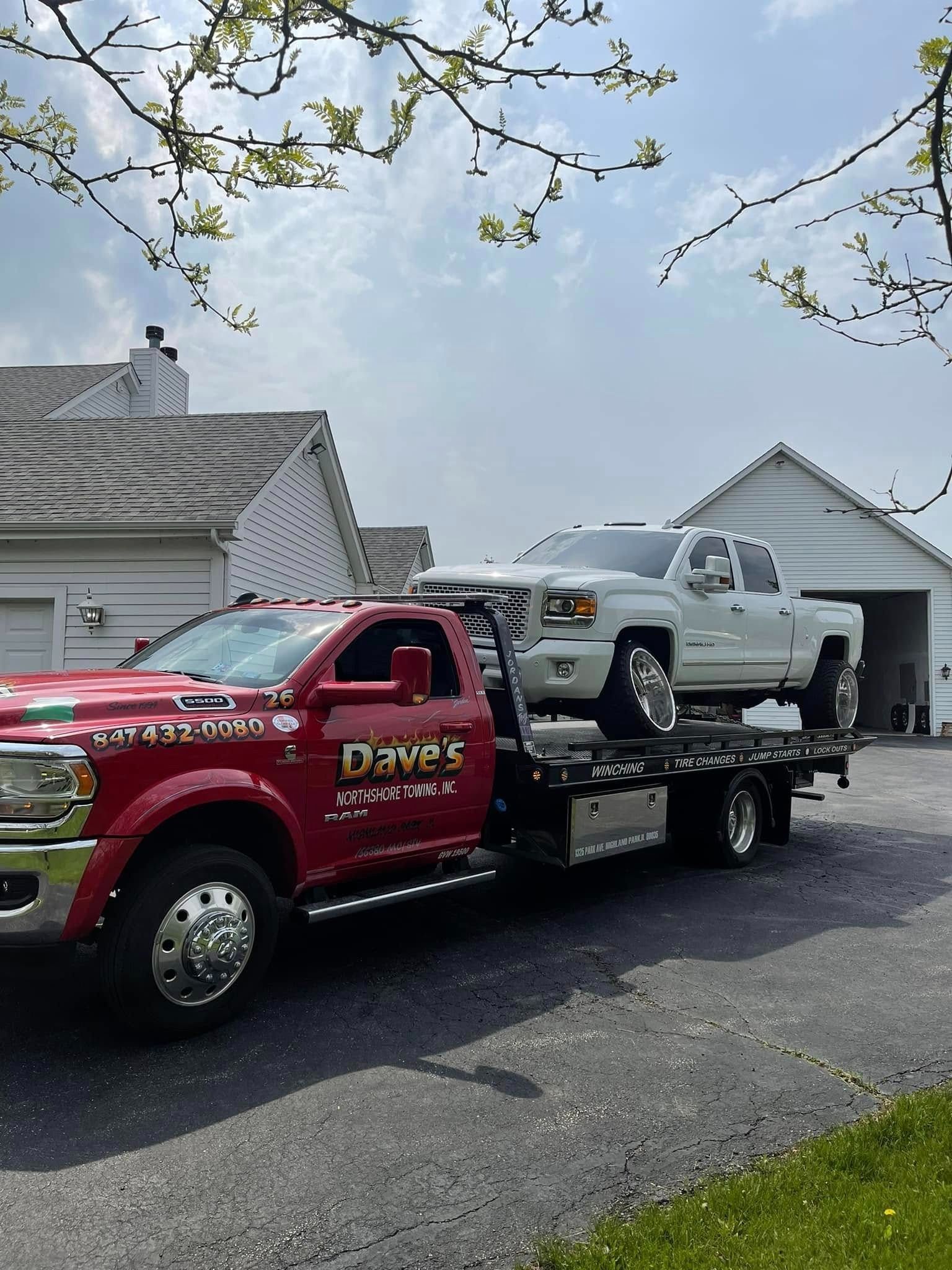 A red tow truck is towing a white truck in a driveway.