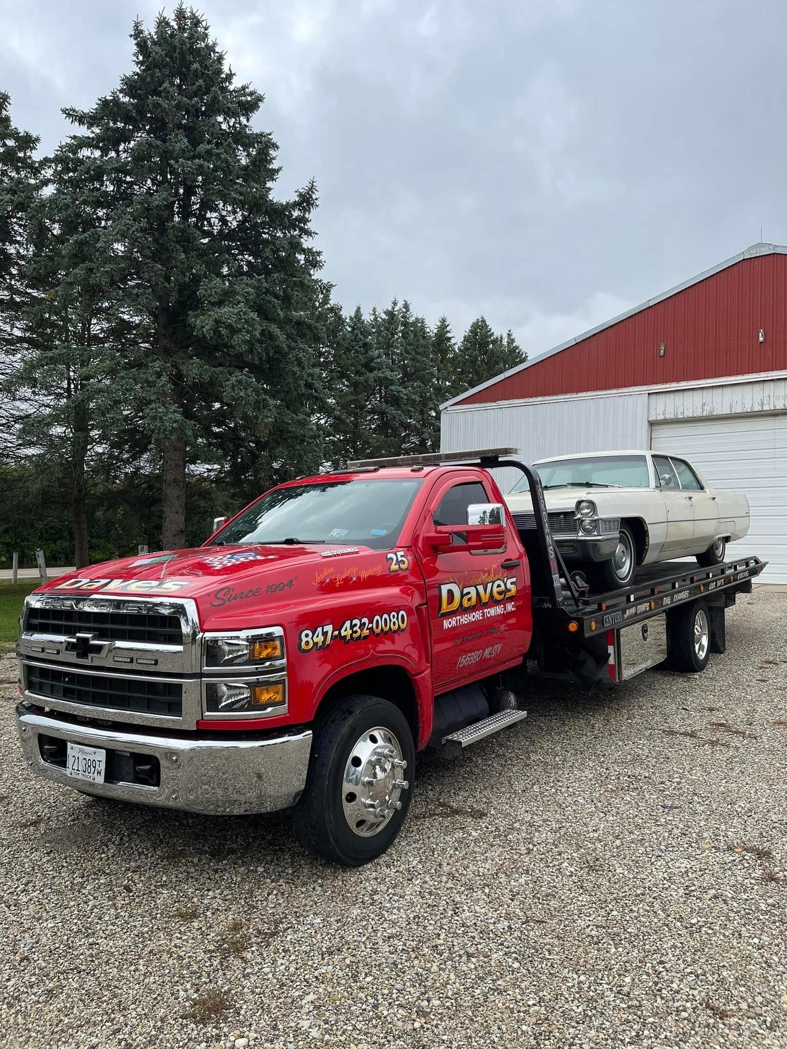A red tow truck is carrying a white car on a gravel road.