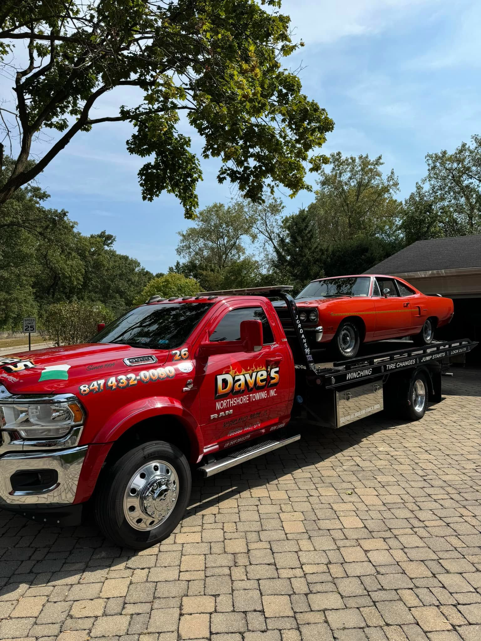 A red tow truck with two cars on the back is parked on a brick driveway.